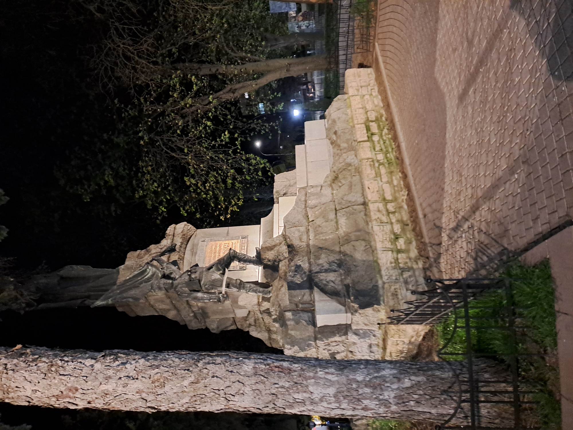 The image shows a monument in the city of Unknown, Unknown. The monument is constructed of large, rough-hewn stones and features a bronze statue of a reclining figure. A bronze plaque is mounted on the monument above the figure. A low, wrought-iron fence surrounds the monument. A brick walkway is visible adjacent to the monument. Trees are in the background, illuminated by streetlights. A tree trunk is prominently visible in the foreground. The scene is dimly lit, suggesting it is nighttime.