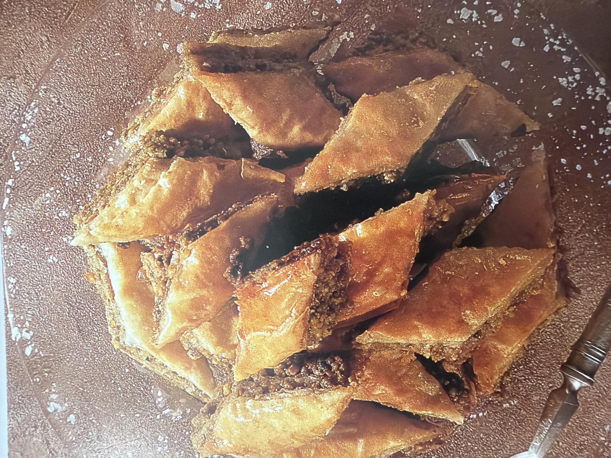 A close-up aerial view showcases a circular arrangement of diamond-shaped baklava pieces, glistening with syrup and sprinkled with coarse sugar crystals against a textured, dark brown background. The baklava, a traditional Mediterranean pastry, is made of thin layers of phyllo dough filled with chopped nuts and bound together with syrup or honey. The pastry exhibits a golden-brown hue, indicating it has been baked to perfection, with some pieces slightly darker around the edges. The syrup coating gives the baklava a glossy finish, reflecting the light and creating highlights.
The arrangement is densely packed, with the individual pieces overlapping and nestled together, forming a visually appealing mosaic. The texture of the background is granular and slightly irregular, possibly a textured platter or serving dish. The scattered sugar crystals add a decorative and textural element, appearing as small, white flecks throughout the scene. A hint of a decorative serving utensil, possibly a fork or knife with an ornate handle, is visible on the right edge of the frame, suggesting that the baklava is prepared for serving or has been recently enjoyed.
There are no people visible in the image. The lighting appears to be bright, highlighting the texture and sheen of the baklava. The overall mood is one of indulgence and sweetness. The image is static, capturing a moment of still life. No text is visible in the media file.