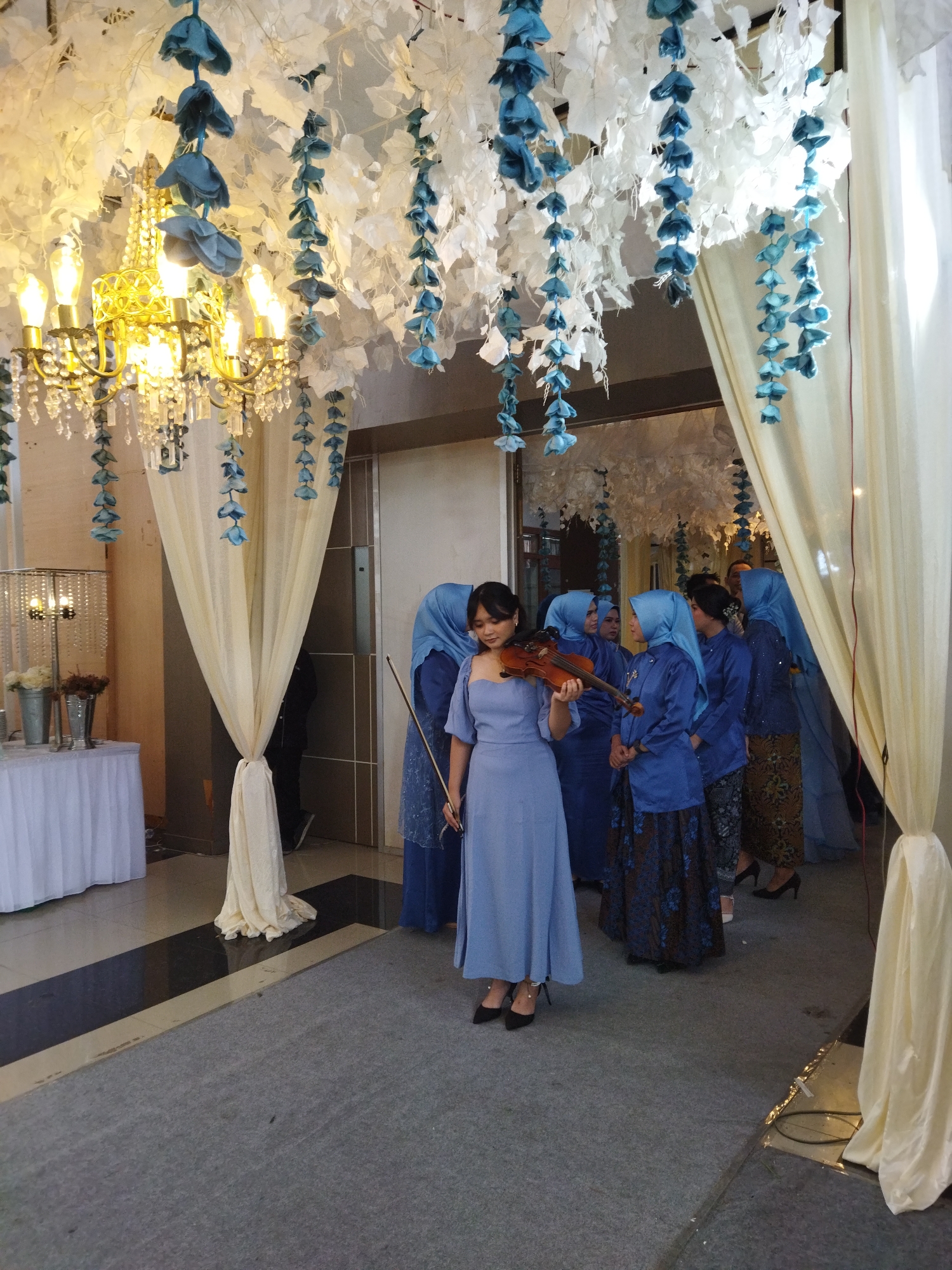In Semarang, Indonesia, a group of women, some wearing blue hijabs and dresses, stand near a doorway draped with sheer cream-colored fabric. A woman in the foreground, dressed in a light blue strapless gown, is playing the violin. She is positioned on a gray carpeted floor. Above, the ceiling is decorated with white artificial foliage and hanging blue floral garlands. A chandelier with multiple bulbs and crystal accents hangs from the ceiling, casting a warm light. To the left, a table covered with a white tablecloth holds decorative items, including buckets with flowers. The overall scene appears to be an indoor event venue.
