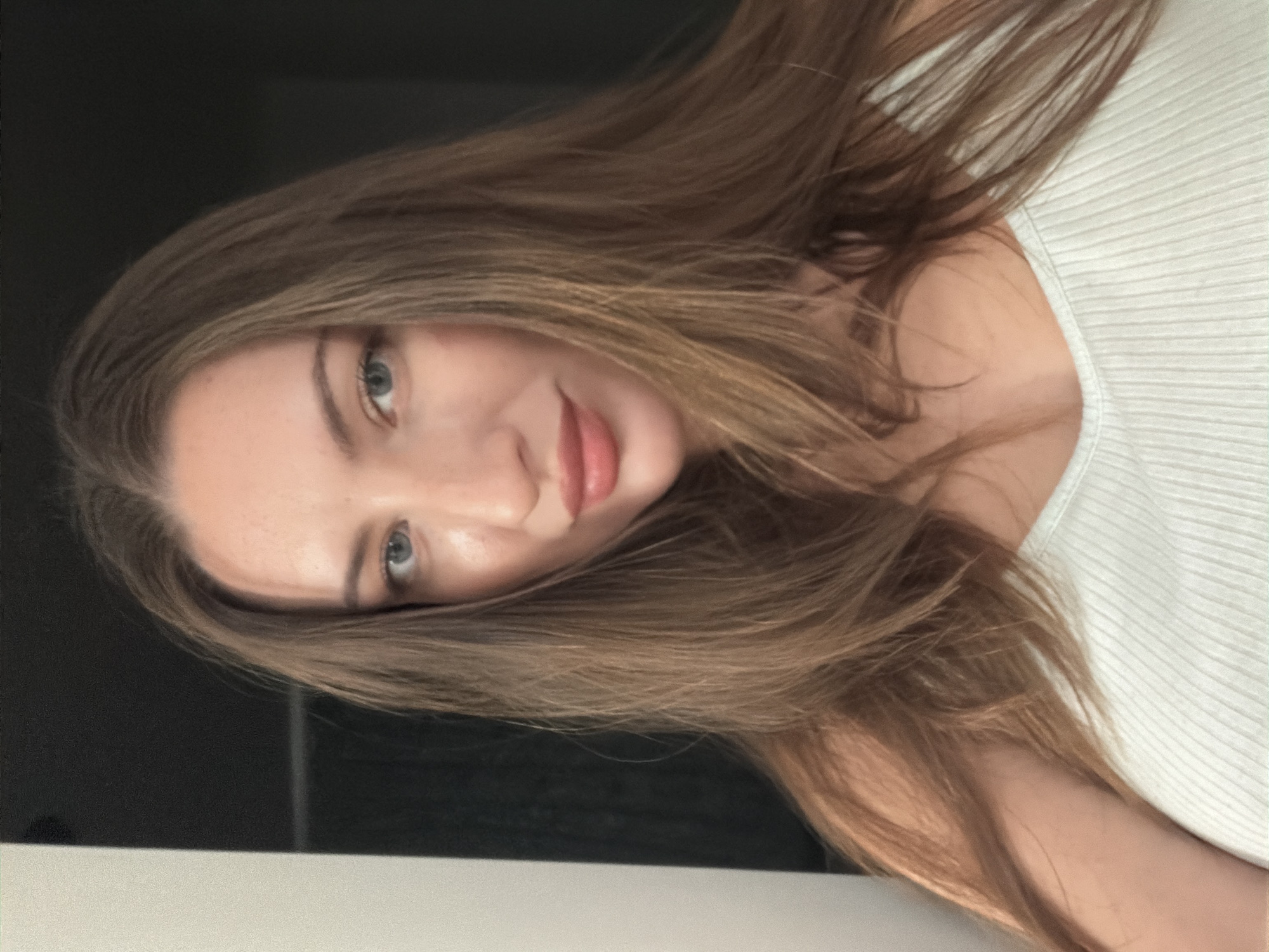 A medium shot shows a woman with long, brown hair and blue eyes. She is wearing a white ribbed tank top. Her hair is parted in the middle and falls around her face and shoulders. She is looking directly at the camera with a slight smile. The background is dark, with a white vertical element on the left side. The lighting suggests an indoor setting. There is no indication of the city of Sharm El Sheikh, Egypt, in the image.
