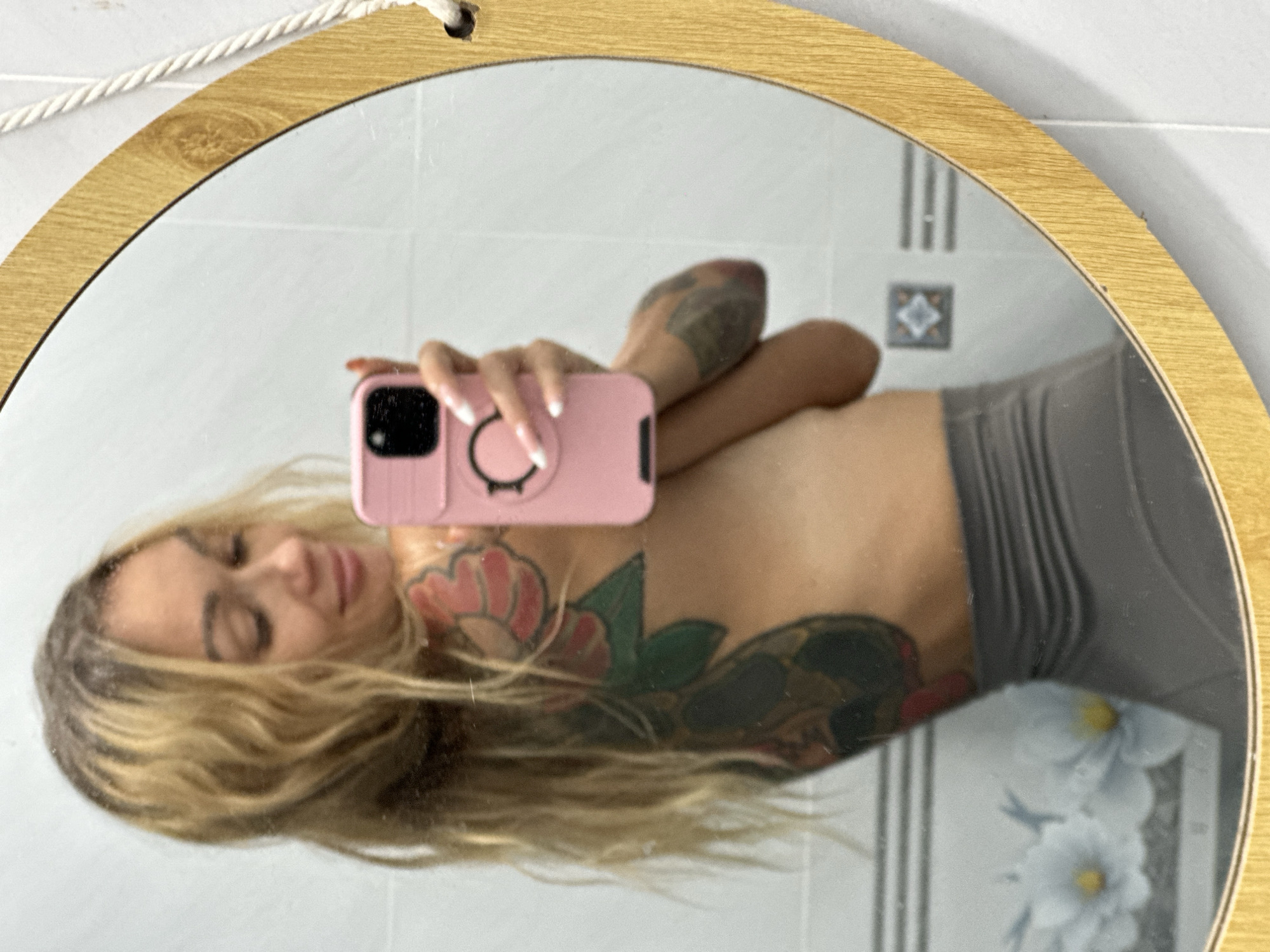 A woman with blonde hair and tattoos on her back and arm is taking a mirror selfie. She is wearing a grey garment. She is holding a pink phone in her right hand, and her left arm is bent, with her hand resting on her hip. The mirror is round and has a wooden frame. In the background, the wall is tiled with a floral pattern visible on the left side. The scene appears to be in a bathroom in Mũi Né, Vietnam.