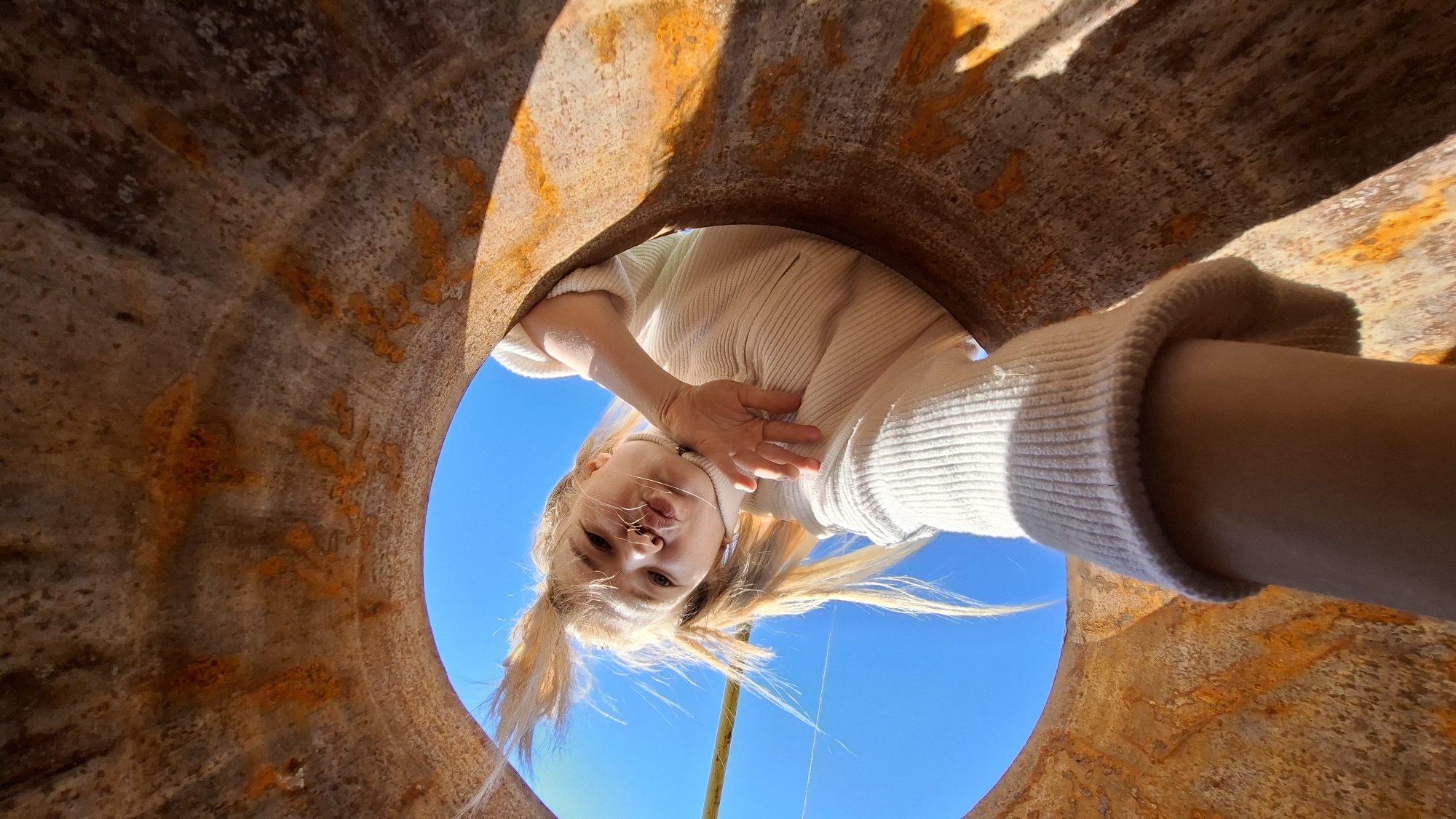 The media file captures a unique low-angle perspective from within a large, heavily rusted metal pipe, looking directly upwards. A young person, likely female, with blonde hair styled in pigtails, is peering down into the pipe opening. She is wearing a light cream or off-white ribbed long-sleeved top, with matching ribbed cuffs visible on her arm. Her expression is playful, featuring a pouting or kissing face directed towards the camera, while one hand is partially raised near her chin.
The background consists of a vibrant, clear blue sky, indicating bright daylight and sunny weather. The inner surface of the pipe displays prominent textures of orange-brown rust and corrosion, creating a raw, industrial aesthetic. The shadows cast by the pipe's rim add depth to the frame. The scene is set in Большой Букор, Russia. No visible text is present.