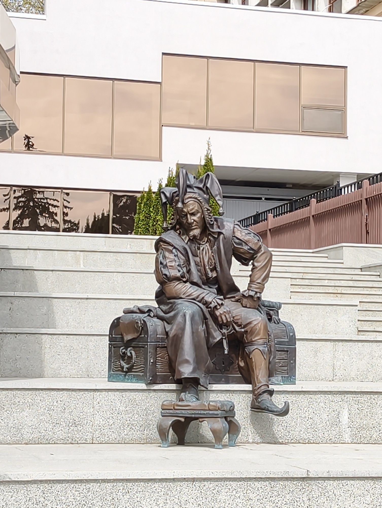 A full shot captures a bronze sculpture of a man dressed as a jester, seated upon a chest. He is positioned on a gray granite staircase with a small stool under his feet. The jester wears a traditional jester's hat and costume, complete with a pendant around his neck and pointed shoes. He holds an object in his hand.
In the background, a modern building with tinted windows reflects trees and sky. A brown metal fence is also visible. The sculpture is located in Saratov, Russia.