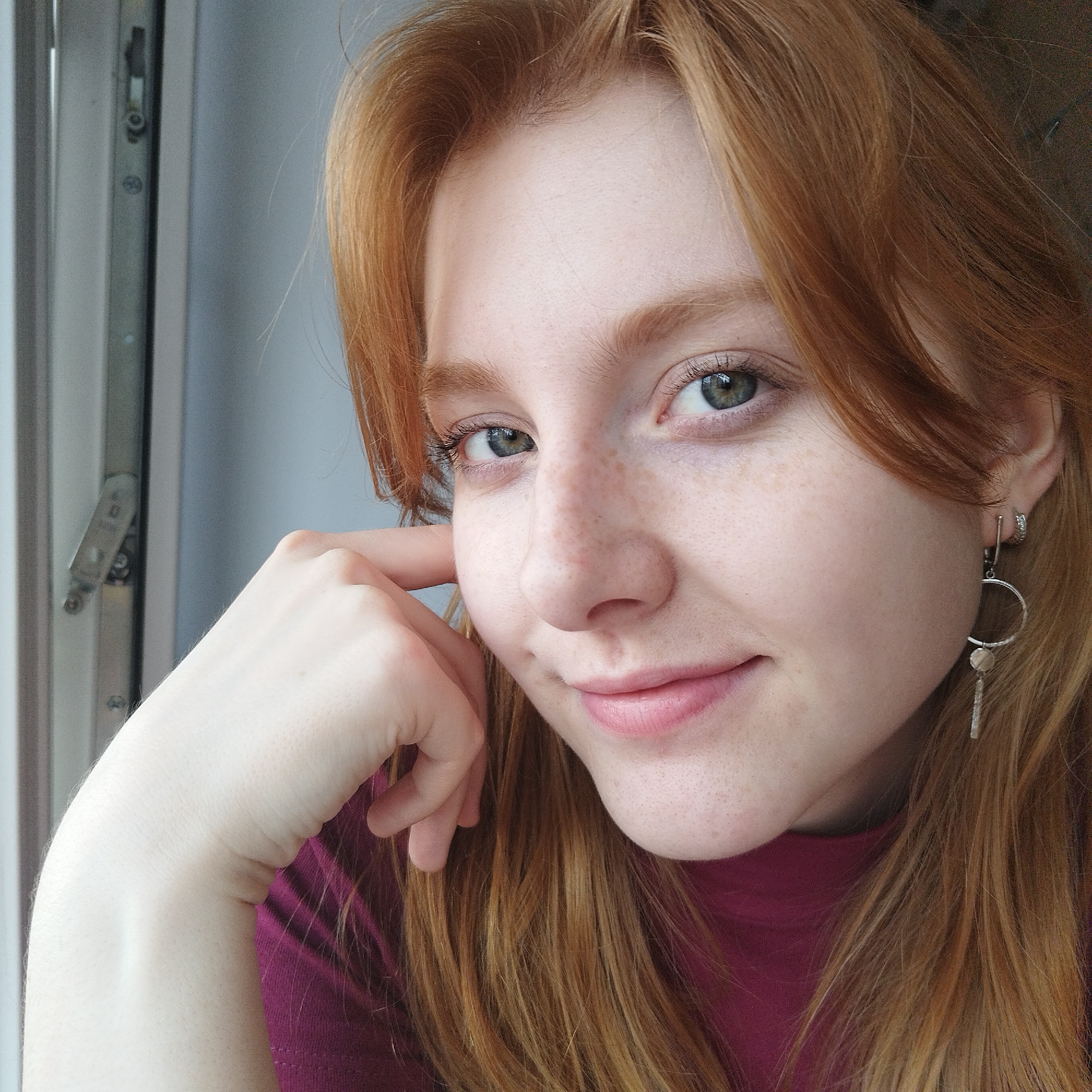 A young woman with fair skin, red hair, and freckles is posing for a close-up portrait. She is smiling subtly with green eyes and wearing a dark pink or maroon-colored shirt. Her right hand is raised, with her fingers gently touching her cheek. She is wearing an earring that has a circle and a bar hanging down. In the background, a window frame is visible on the left side of the frame. The image does not provide enough information to determine the exact location of the woman, so the specific location in the city of Yekaterinburg, Russia, cannot be pinpointed.
