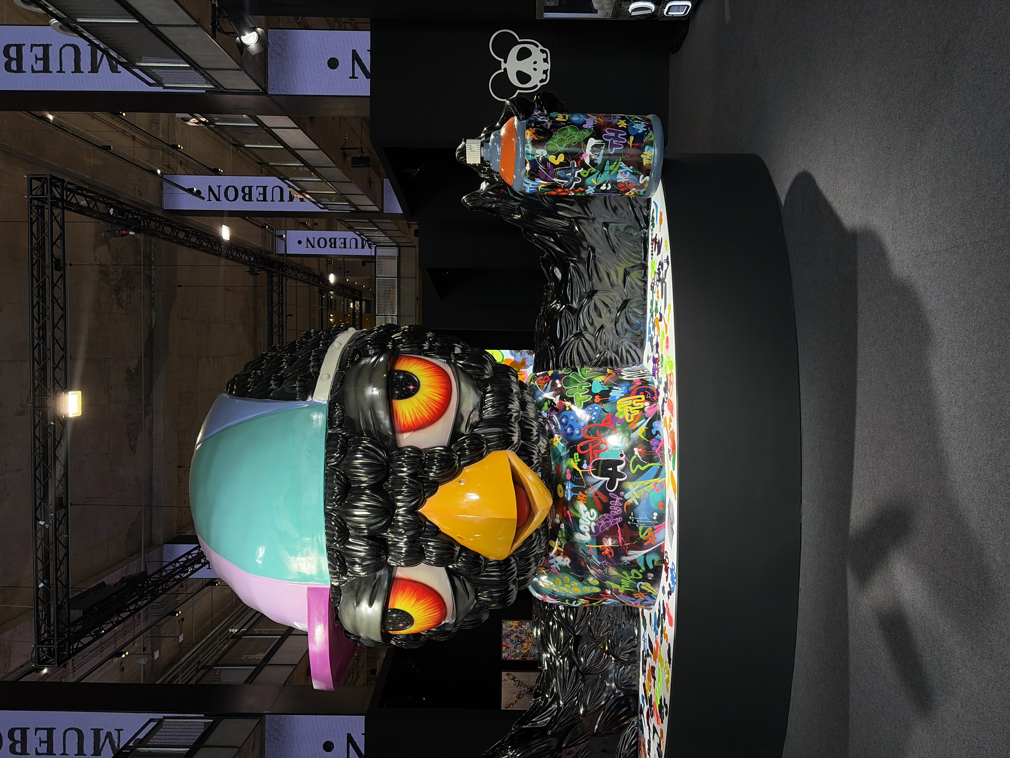 A large, surreal sculpture stands within an interior space, possibly a gallery or convention center, in Bangkok, Thailand. The sculpture, featuring a cartoonish head resembling a bird or penguin, is adorned with vibrant, colorful graffiti and spray-painted designs. The figure's head is multi-colored, incorporating shades of blue, pink, and purple, with large, stylized eyes that have orange irises. Its beak is rendered in a bright yellow. The sculpture appears to be holding a spray paint can, also decorated with graffiti.
The background features signs with the word "MUEBON" illuminated in white, suggesting a brand or exhibition. The architectural structure is industrial, with metal beams and grid-like patterns visible. The floor is carpeted in a dark grey or black, contrasting with the bright colors of the sculpture. The lighting is a mix of natural and artificial, illuminating the sculpture and the surrounding environment. The photograph is taken at an angle, giving a unique perspective of the artwork and the space it occupies.