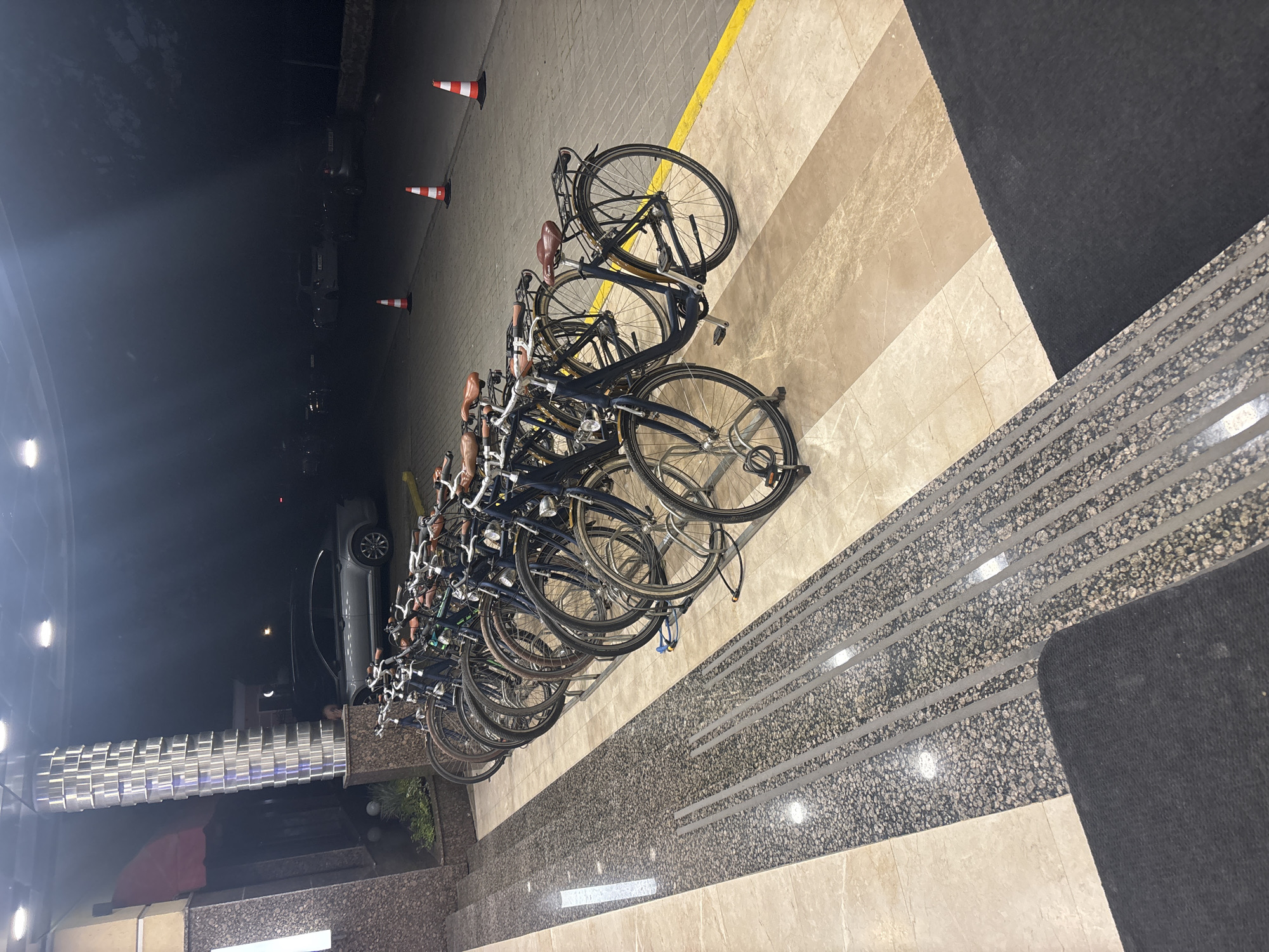 The image depicts a row of bicycles parked on a tiled and striped surface next to a dark-colored rug and tactile paving. In the background, there is a street with parked cars and orange traffic cones. The scene appears to be at night or in low light conditions. Given the presence of tactile paving and the general urban look, it is likely a public space or the entrance area of a building. The image does not contain any explicit indicators of the location being Plovdiv, Bulgaria, but this can not be ruled out.