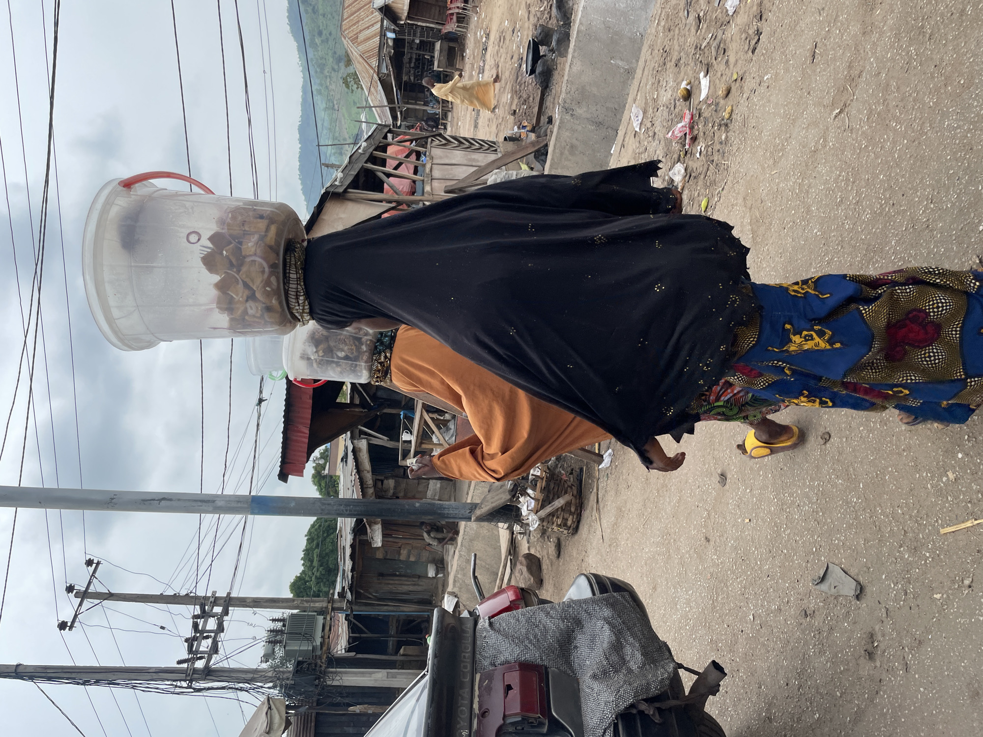 In Lokoja, Nigeria, two women are seen walking away from the camera. The woman in the foreground is wearing a black hijab and a colorful patterned skirt, carrying a large transparent plastic bucket filled with food on her head. The woman behind her is wearing an orange top and a patterned skirt, also carrying a bucket on her head. They are walking on a dirt road in what appears to be a market area, with makeshift stalls and buildings on either side. In the background, there are power lines and hills. A portion of a dark-colored car is visible on the left side of the image. The overall scene depicts a busy street scene in a Nigerian city.