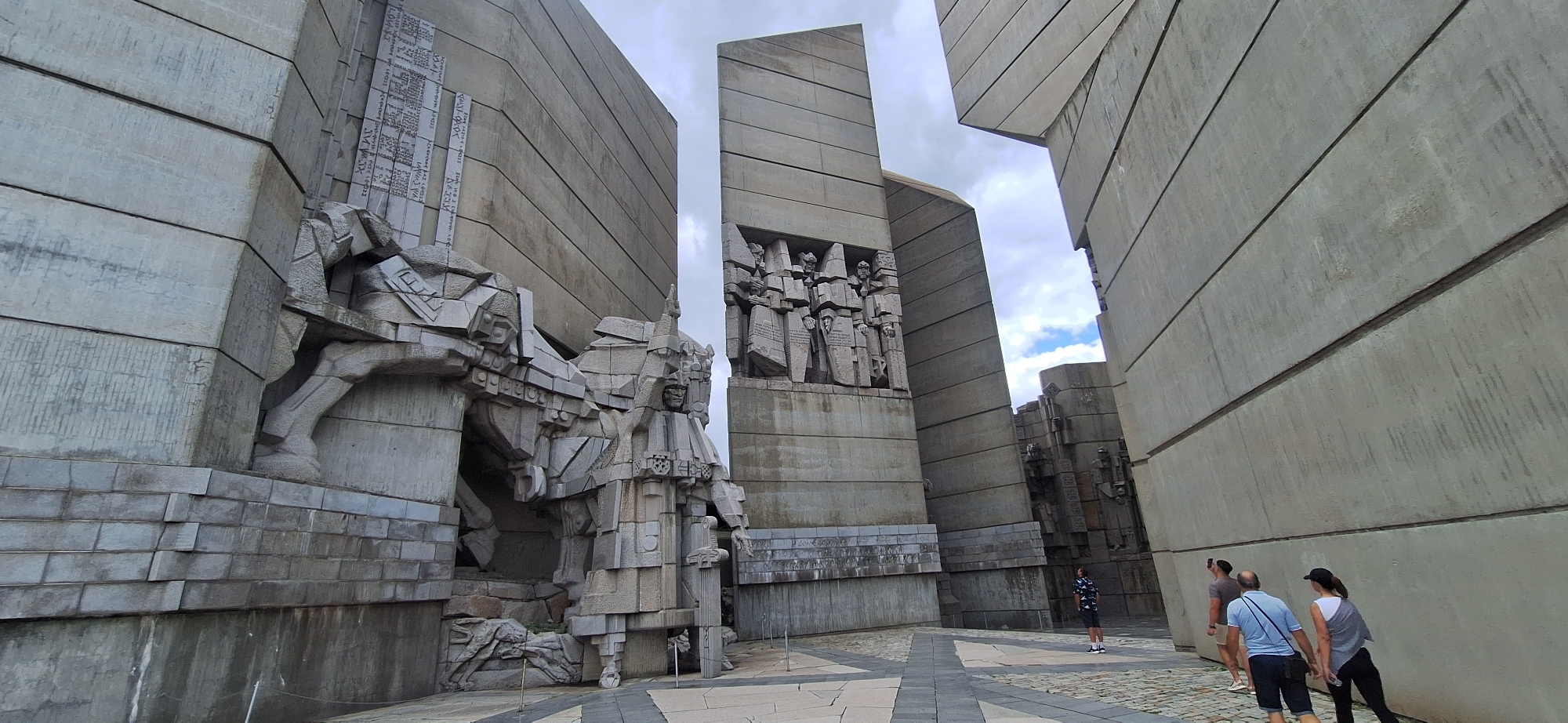 The image shows the Monument to the Bulgarian Soviet Friendship, a monumental Soviet-era structure located in Shumen, Bulgaria. The monument is made of concrete and features large, abstract sculptures and reliefs. In the foreground, several people are walking and observing the monument. The sky is overcast with patches of blue. The paving stones in front of the monument are laid in a geometric pattern.