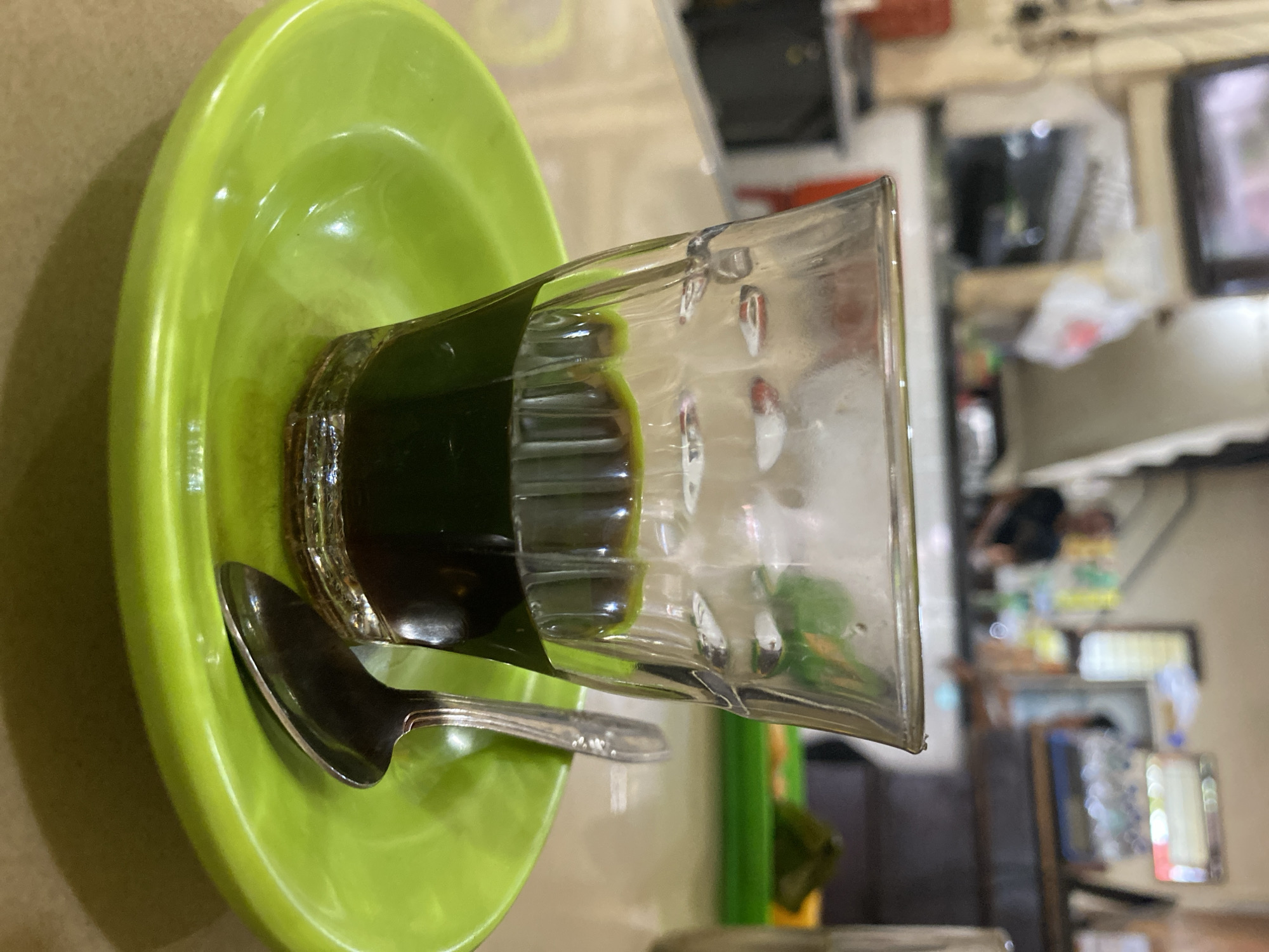 A close-up, medium shot captures a glass of dark liquid, possibly coffee, resting on a bright green saucer. A silver spoon lies on the saucer to the right of the glass. The glass is short and stout, with vertical ridges near the base, and contains a dark, opaque liquid with a visible greenish tint on the surface. The saucer is slightly curved, showcasing its vibrant green color and glossy finish. The background is blurred, suggesting an indoor setting with ambient lighting. Hints of a counter and shelves are visible, along with indistinct figures of people, suggesting a public space like a cafe or warung. The overall impression is of a simple, everyday moment captured in Lhokseumawe, Indonesia, a city known for its culinary scene and bustling local eateries.