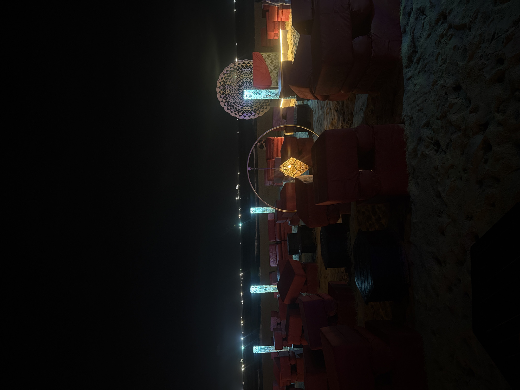 The image shows a nighttime scene in an unknown location. Soft, red cushions are stacked in seating arrangements on a sandy surface, possibly a beach. Illuminated decorative objects and string lights enhance the ambiance. The presence of lights suggests an event or designed space, although the nature of the event or space cannot be determined with certainty.