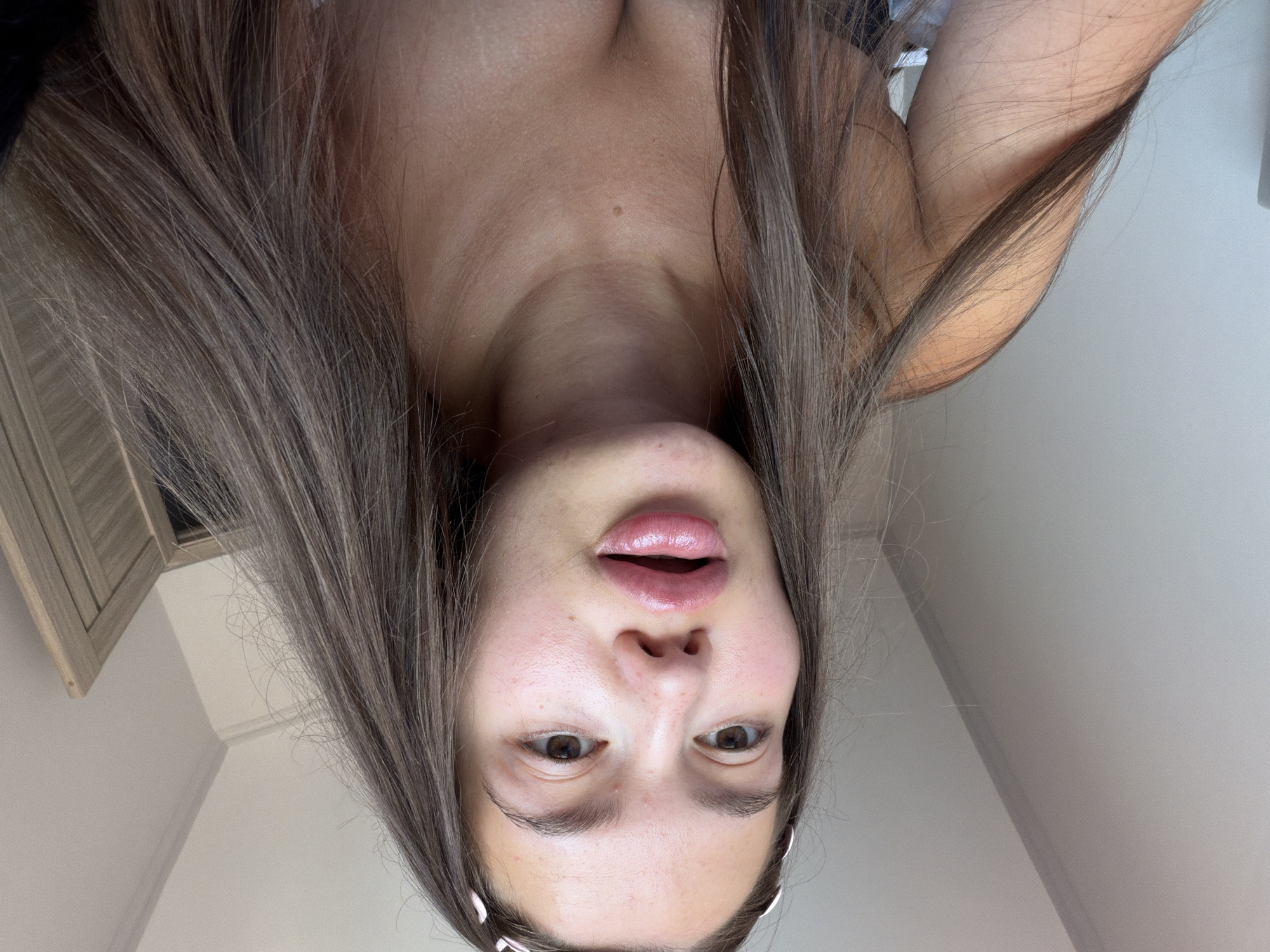 A young woman with long, brown hair and light skin is captured in a close-up, low-angle selfie. She is wearing a dark top, and her hair is falling over her shoulders. Her lips are full and glossy, and she has light pink blush on her cheeks. The background is a light-colored wall, with a subtle shadow line suggesting a corner and a wooden door or cabinet to the right. The scene appears to be indoors, possibly in a bedroom or living space. There is no indication of the location being in Sirius, Russia.