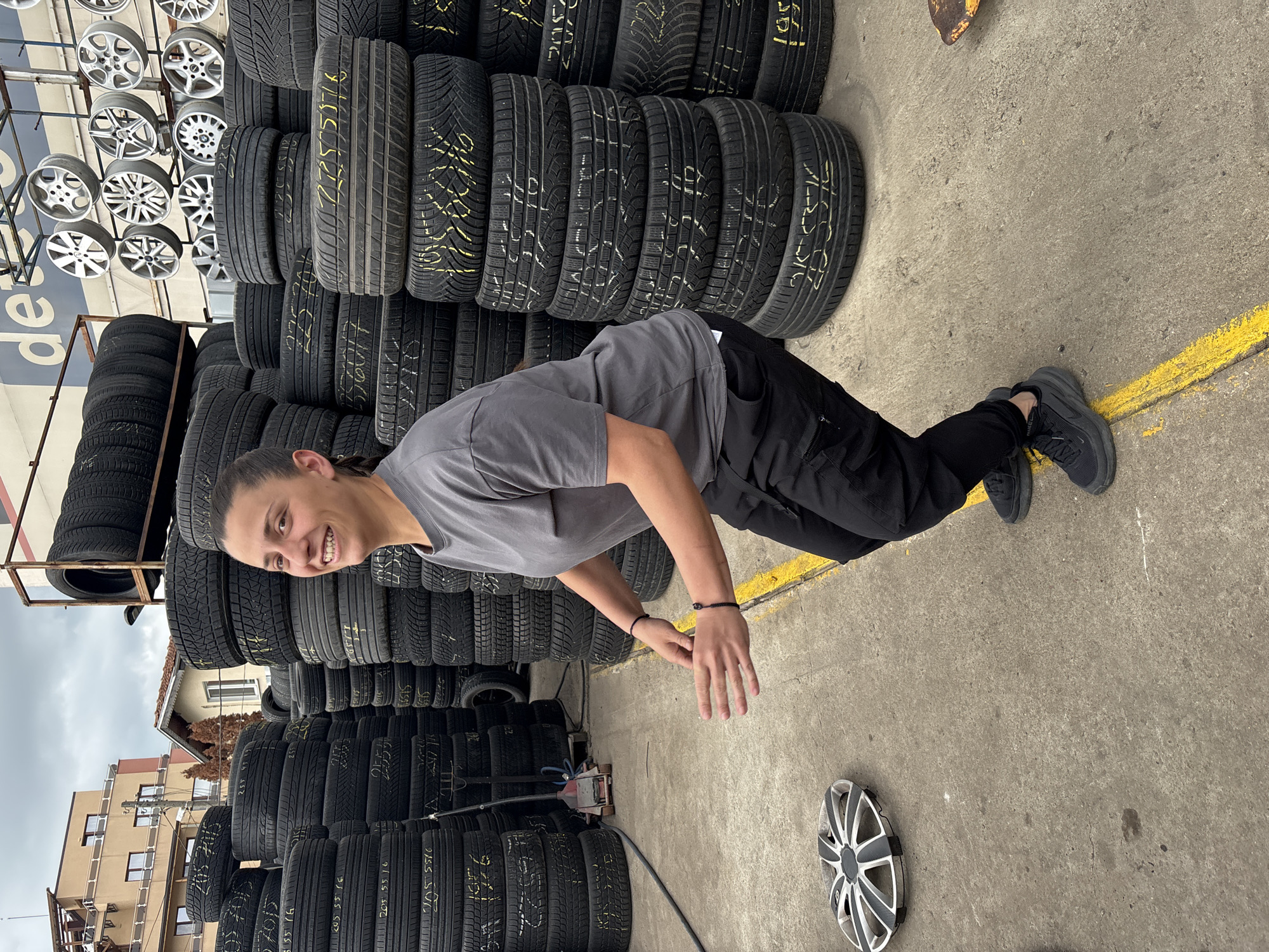 A young person with their hair tied back is crouching in front of a large stack of used car tires. The tires are stacked in rows and have yellow markings on them. There are wheels displayed on the building behind the tires. The location is likely a tire shop in Sofia, Bulgaria, based on the Cyrillic script on the tires and the urban environment. The person is wearing a gray t-shirt, black pants, and black shoes. They are smiling and looking at the camera. A hubcap sits on the ground beside the yellow painted line. A hydraulic jack is present near the tires. Buildings are visible in the background.