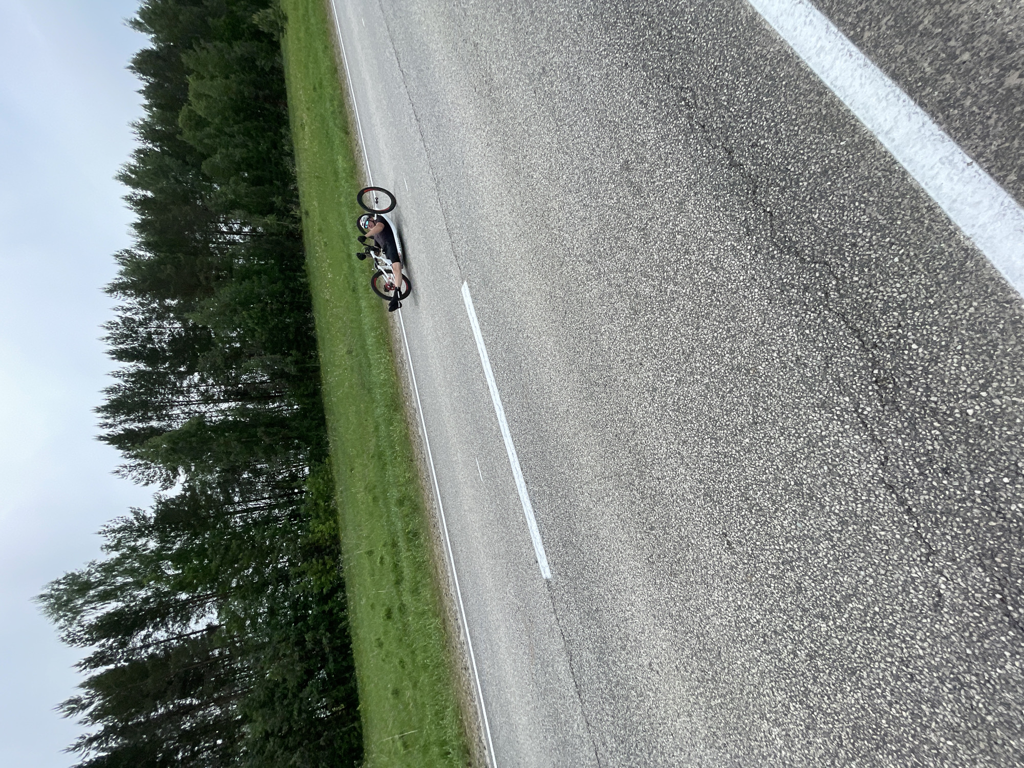 The image shows a recumbent bicycle being ridden on a paved road in Перевозное, Russia. The road has white lane markings, and a grassy area bordered by trees runs alongside it. The sky appears overcast. The bicycle is being ridden by an individual wearing what appears to be a helmet and dark clothing.