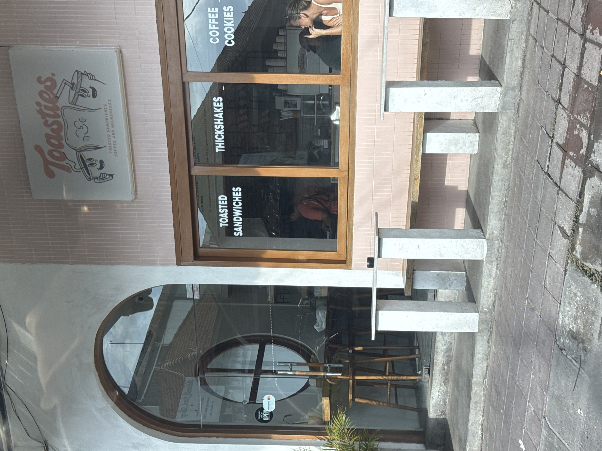 This image captures the exterior of a modern cafe named "Toasties." in North Kuta, Indonesia, on a sunny day. The cafe features a light pink tiled upper facade and a lighter, possibly white or pale grey, lower wall. A large rectangular window and an arched glass door dominate the front.
Above the rectangular window, a vintage-style sign with an illustration of two animated cups/mugs holding a toasted sandwich proudly displays the cafe's name, "Toasties.", along with the offerings "TOASTED SANDWICHES COFFEE AND MILKSHAKES." Inside the window, menu items are advertised vertically: "COFFEE," "COOKIES," "THICKSHAKES," and "TOASTED SANDWICHES." Partial text for sandwich fillings like "TOMATO, BASIL PESTO, CHEESES" and pricing information "All prices In" are also visible.
Through the upper part of the rectangular window, a woman with light hair is seen leaning over a counter, with another person partially obscured behind the counter. Inside the cafe, through both the main door and the lower part of the window, wooden furniture and the reflection of a person in a reddish-orange shirt are visible. The glass door has "Come in," "OPEN," and "Welcome" stickers.
In front of the cafe, an outdoor seating area is set up with stark, light-colored concrete block benches and tables, casting clear shadows on the building, indicating bright daylight. The ground is paved with square and rectangular cobblestones. The overall scene suggests a vibrant, casual dining spot open for business.