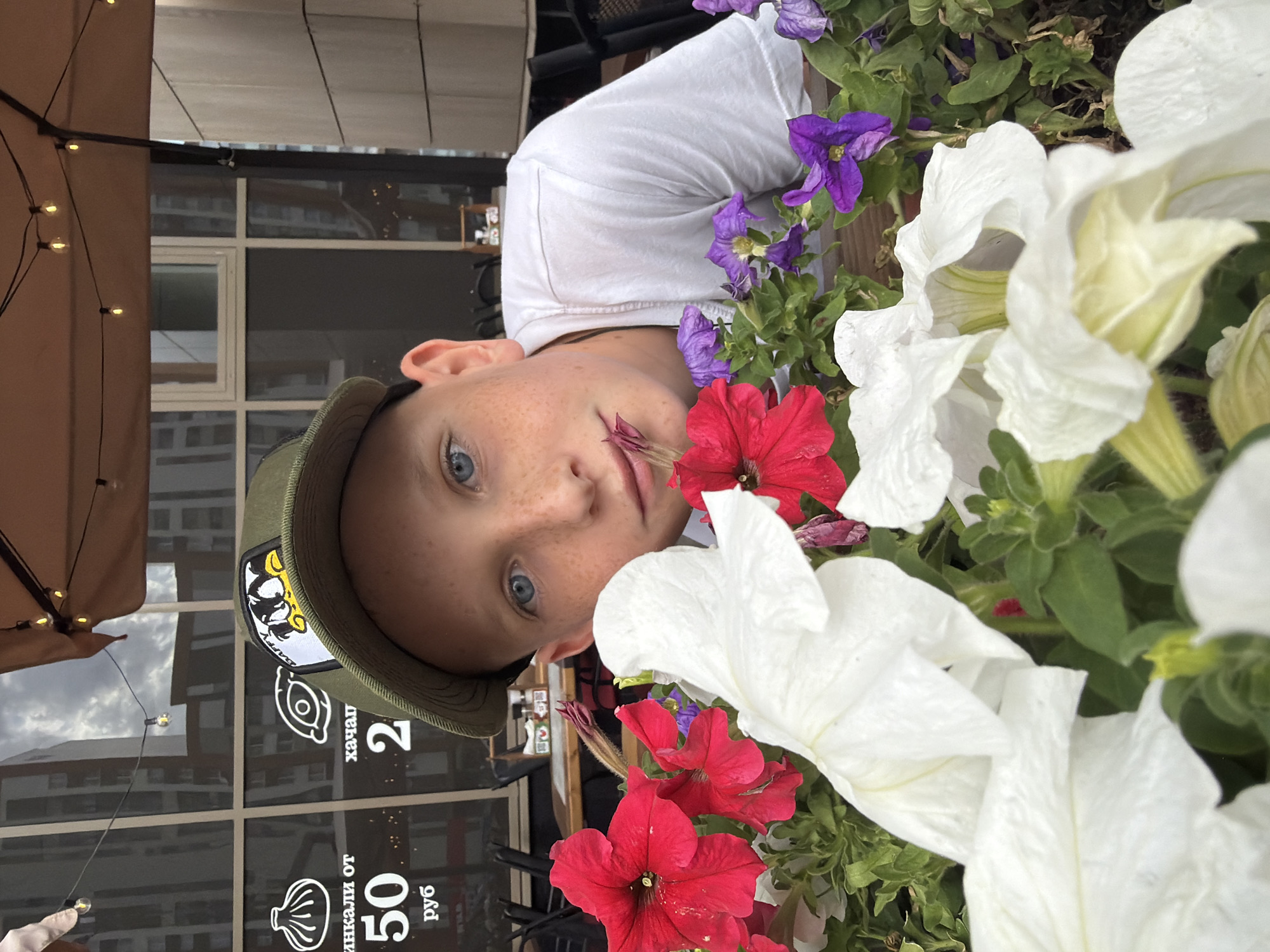 A young boy with freckles and blue eyes, wearing a green baseball cap with a cartoon patch, looks towards the camera from behind a planter filled with red, white, and purple petunias. He is wearing a white t-shirt. The background shows a glass window with building reflections and outdoor cafe seating. Text on the window indicates prices for "khinkali" and "khachapuri," suggesting the location is a cafe or restaurant in Yekaterinburg, Russia. Specifically, the text visible on the window translates to "Khinkali from 50 rubles" and "Khachapuri 2."