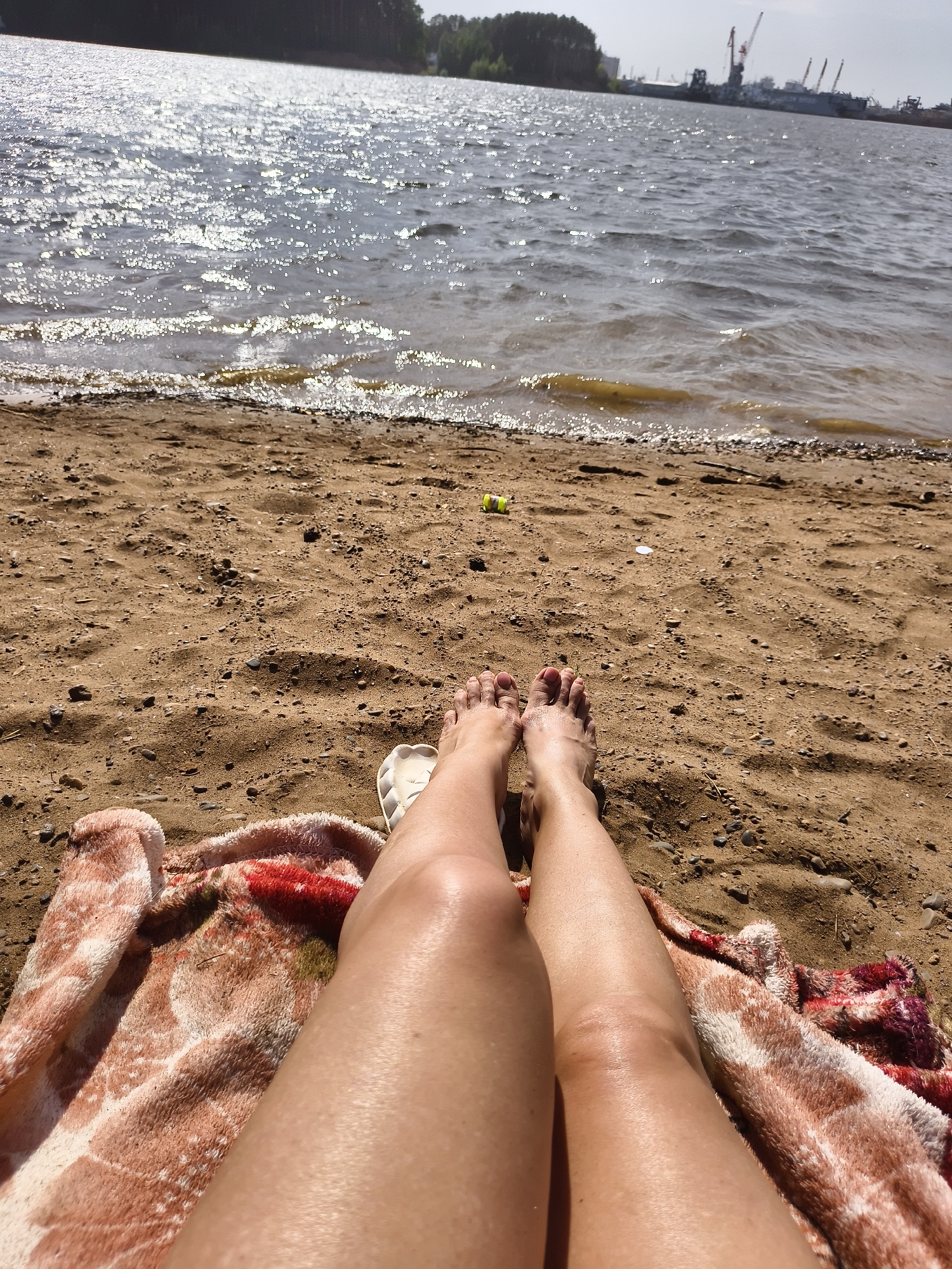 A sunny beach scene with a person's legs and feet visible in the foreground. The person is lying on a colorful patterned blanket, with their feet pointing towards the top of the image. The beach is sandy with scattered pebbles. In the background, there is a body of water with sunlight reflecting off its surface. Across the water, there is a shoreline with trees and some industrial structures, including cranes. The location is in the city of Unknown, Unknown.