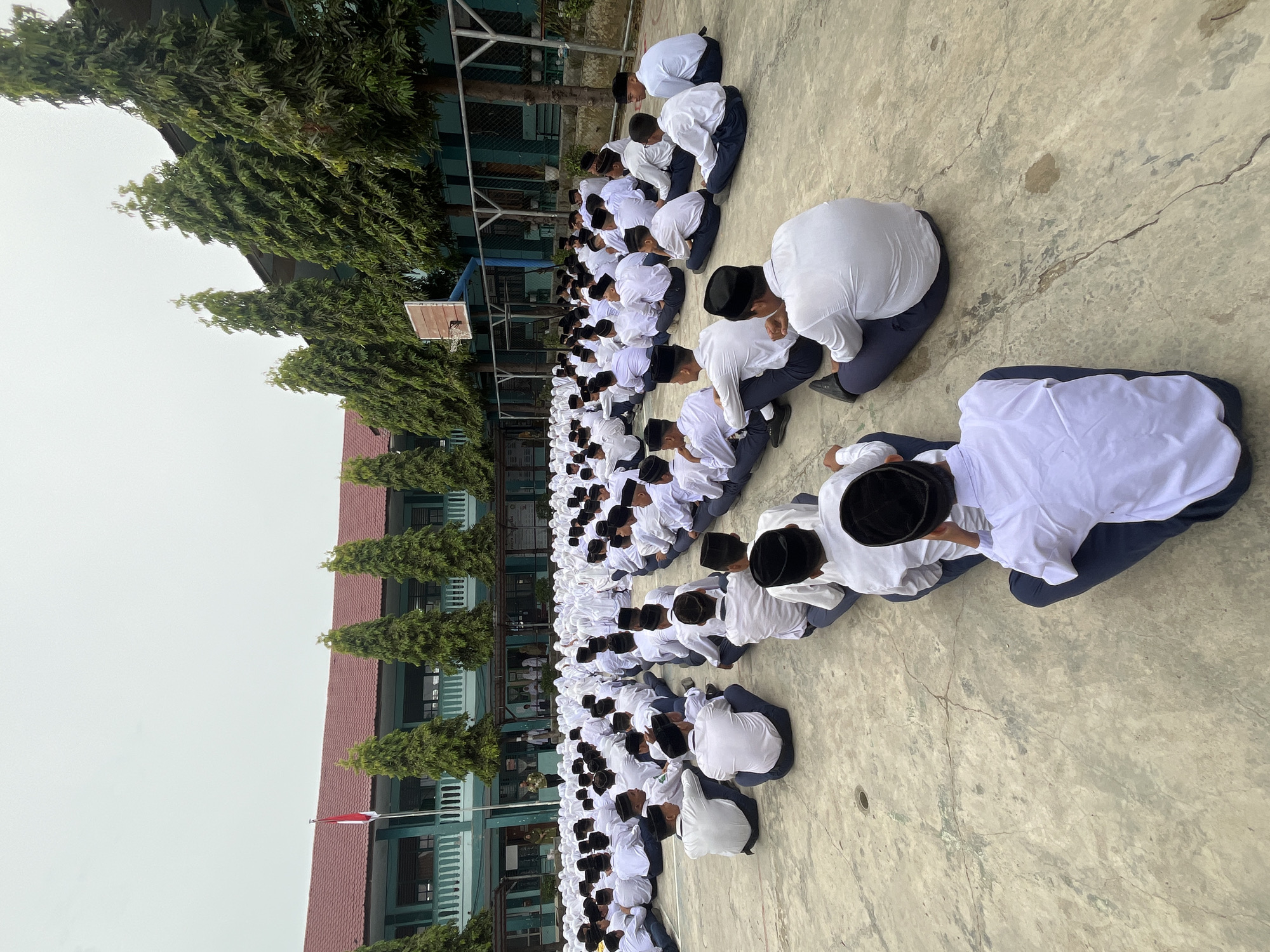 The image depicts a large gathering of male students seated on a concrete ground in what appears to be a school courtyard in Lhokseumawe, Indonesia. The students are uniformly dressed in white long-sleeved shirts, dark blue trousers, and black peci hats, indicating they are likely participating in a school assembly or religious activity. The environment features a light-colored concrete floor with some cracks and stains, suggesting it's an outdoor space. In the background, a multi-story building with a red tiled roof and light green walls stands, possibly the school building. Tall, slender green trees line the side of the building, and a basketball hoop with a wooden backboard is visible. The sky is overcast, suggesting a cloudy day. The students are mostly facing away from the camera, with some individuals in the foreground looking towards a person or group standing at a distance, perhaps a teacher or speaker. The overall scene is orderly, with the students arranged in rows.