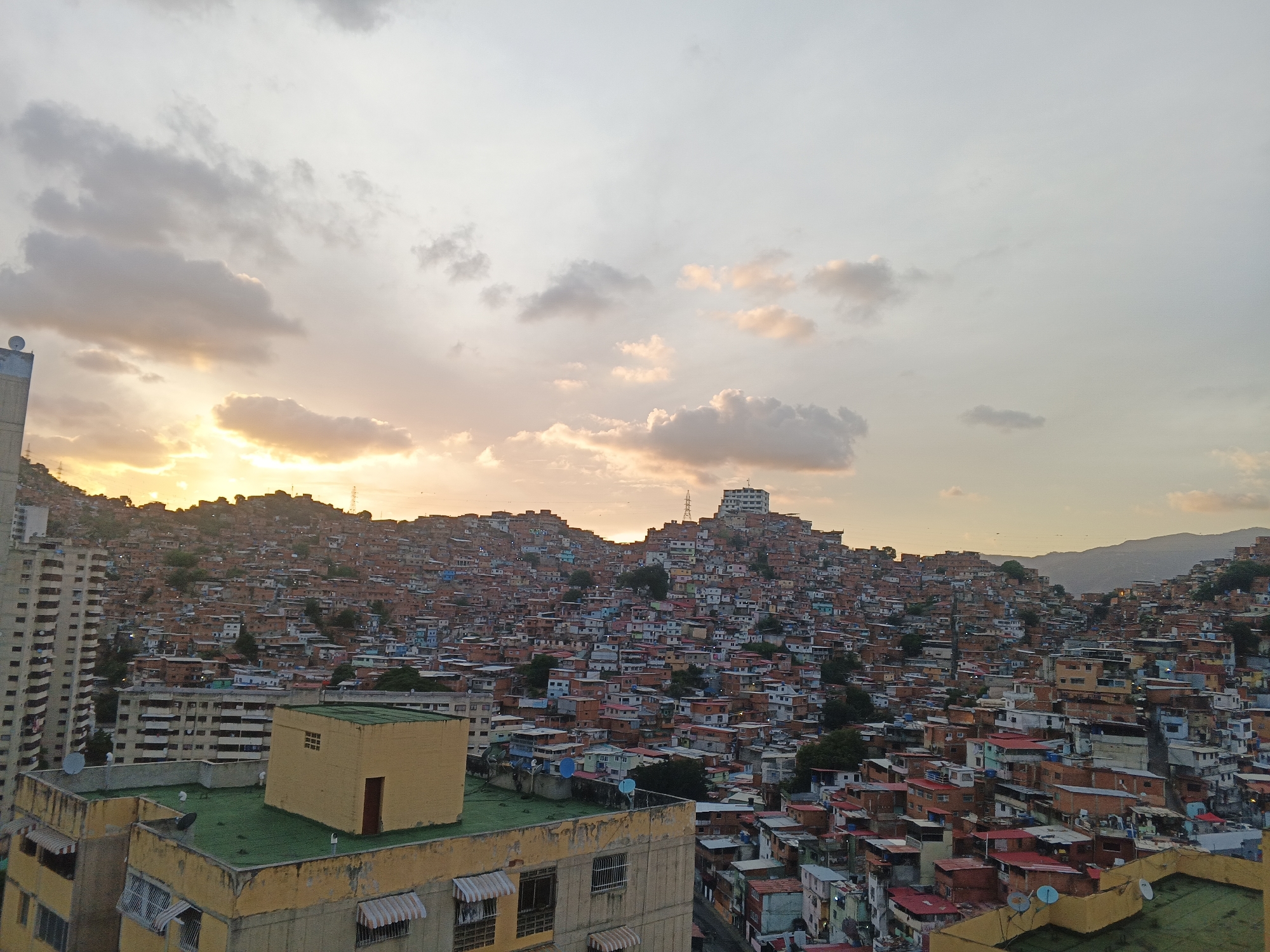 This image presents an expansive urban landscape in Caracas, Venezuela, captured during the golden hour, indicating either sunrise or sunset with a warm, bright light source near the horizon on the left. The foreground features a cluster of urban buildings, including a prominent yellow structure with a flat green roof, dark brown door, and small window on its roof, along with striped awnings over ground-level windows. To its left, a taller, beige multi-story building with numerous balconies rises. Multiple white satellite dishes are visible on the rooftops of these foreground structures.
The midground and background are dominated by a vast informal settlement, locally known as a barrio, which sprawls across multiple steep hillsides. These hills are densely populated with countless small, multi-colored houses, primarily constructed from brick and concrete, featuring a variety of roof colors including red, orange, blue, green, and white. The architecture shows extreme density, with structures appearing to be built one upon another up the slopes. Interspersed sparingly among the houses are trees and sparse vegetation. A taller, white, multi-story building with numerous windows stands conspicuously atop one of the highest hills in the distance. A power transmission pylon is visible on a ridge to the left of this white building. The sky above is partially cloudy, with scattered cumulus clouds that reflect the warm light with shades of orange and pink near the horizon, transitioning to grayer and lighter blue hues higher up. No people are discernible in the scene.