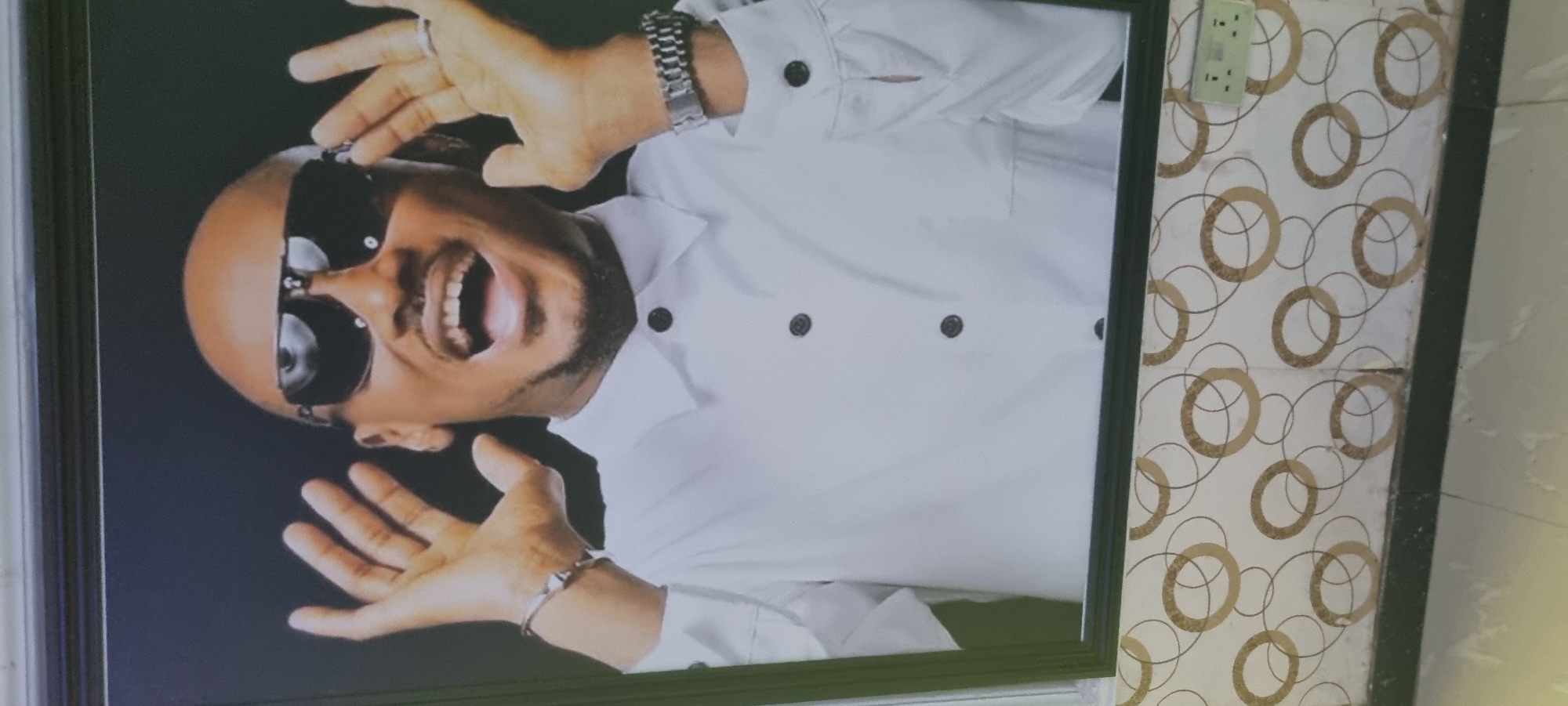 A framed picture of a bald man wearing sunglasses and a white shirt hangs on a wall with a circular pattern. The picture is rotated slightly clockwise. The man is smiling and holding his hands up. A plug socket is visible above the picture frame. The location is Alasia, Nigeria.