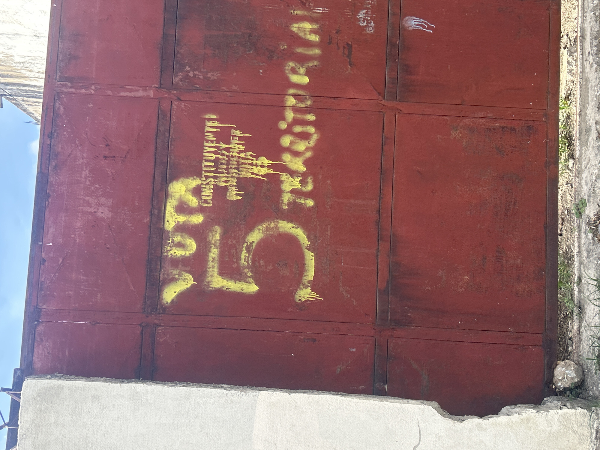 The image shows a weathered red metal gate with yellow spray paint graffiti. The graffiti reads "VOTA 5 CONSTITUYENTE TERRITORIAL". The gate is divided into four rectangular panels by horizontal and vertical supports. The left panel of the gate has a portion of a white concrete structure attached to its left side. The graffiti is applied in a stenciled manner, with some areas showing drips. The "5" is particularly prominent and large, painted in yellow with drips. Above the "5" and to its right, the word "VOTA" is also visible in yellow. To the right of "VOTA", the word "CONSTITUYENTE" is partially visible with some dripping paint. Below "CONSTITUYENTE" and partially obscured by drips, some smaller text is present, which is difficult to decipher but appears to be "RECLAMA". The word "TERRITORIAL" is painted in yellow on the lower right panel of the gate, also with some drips. The overall condition of the gate suggests it is old and exposed to the elements, with rust visible in some areas. The background shows a bright blue sky with some white clouds. The ground in front of the gate is covered with sparse green grass and some gravel. The image appears to be taken outdoors, likely in an urban or semi-urban environment in Santa Ana, Venezuela, given the context of the graffiti.