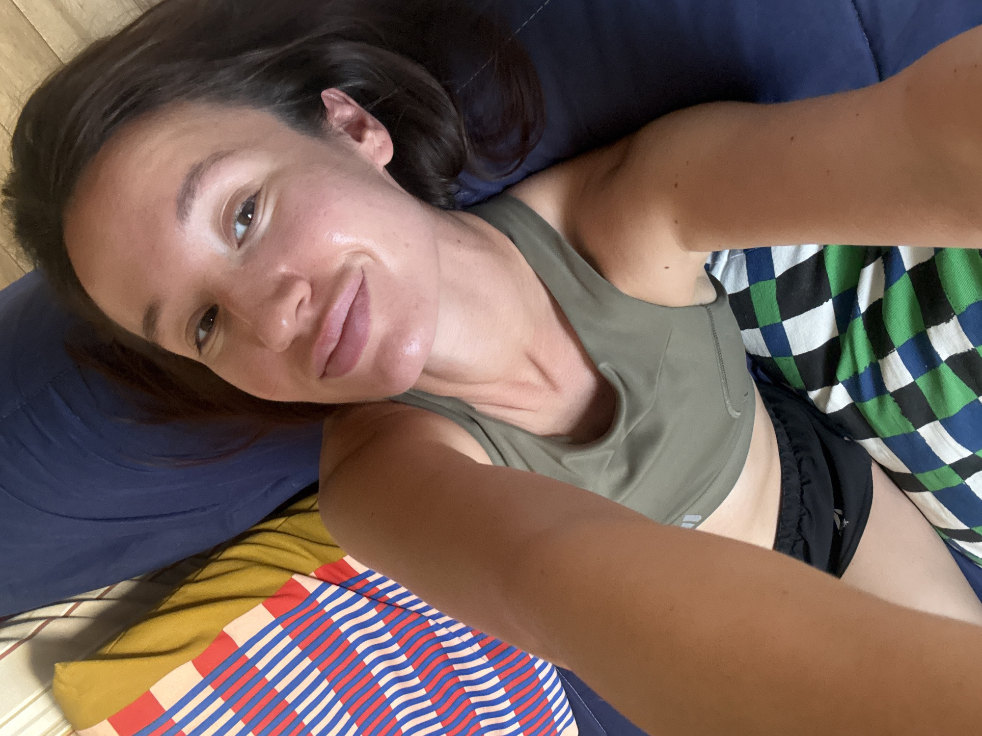 A young woman with dark hair and fair skin is captured in a selfie, lying down indoors. She has a relaxed, subtle smile and her brown eyes are looking directly at the camera. Her skin appears dewy. She is wearing a khaki green sports bra or athletic top and black shorts.
She is resting on a dark blue pillow or cushion. To her right, a blanket or fabric with a blue, green, and white checkered pattern is visible. To her left, another patterned fabric with red, blue, and white stripes is present, possibly layered over a yellow fabric. A dark blue fabric also forms part of the background, along with a hint of a light wooden surface in the upper left. The setting appears to be a comfortable indoor environment, suggesting a bedroom or living area. No text is visible in the image. The location context provided is Tel-Aviv, Israel.
