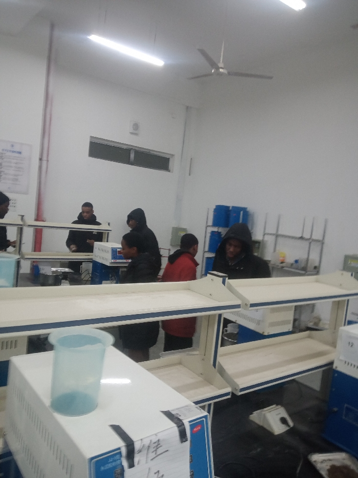 A laboratory setting includes multiple individuals, some in hoodies, working at lab benches. White equipment sits on the benches, and clear plastic containers are visible. In the background, there's a window, a list on the wall, and shelves containing blue containers. The room has a ceiling fan and fluorescent lighting. Overall, the scene depicts people involved in some form of laboratory work or experiment.