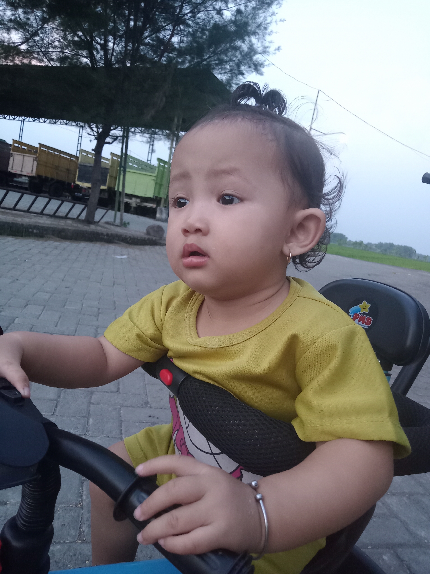 A young child is sitting on a tricycle, with their hands on the handlebars. They are wearing a yellow shirt and a silver bracelet. The child has dark hair tied up in a small ponytail. They are looking off to the side with a slightly pensive expression. The tricycle has a black seat and a blue frame. The background shows a paved area and an open space with a grassy field. There are some trucks parked under a covered structure. The photo was taken in Bungur, Indonesia.