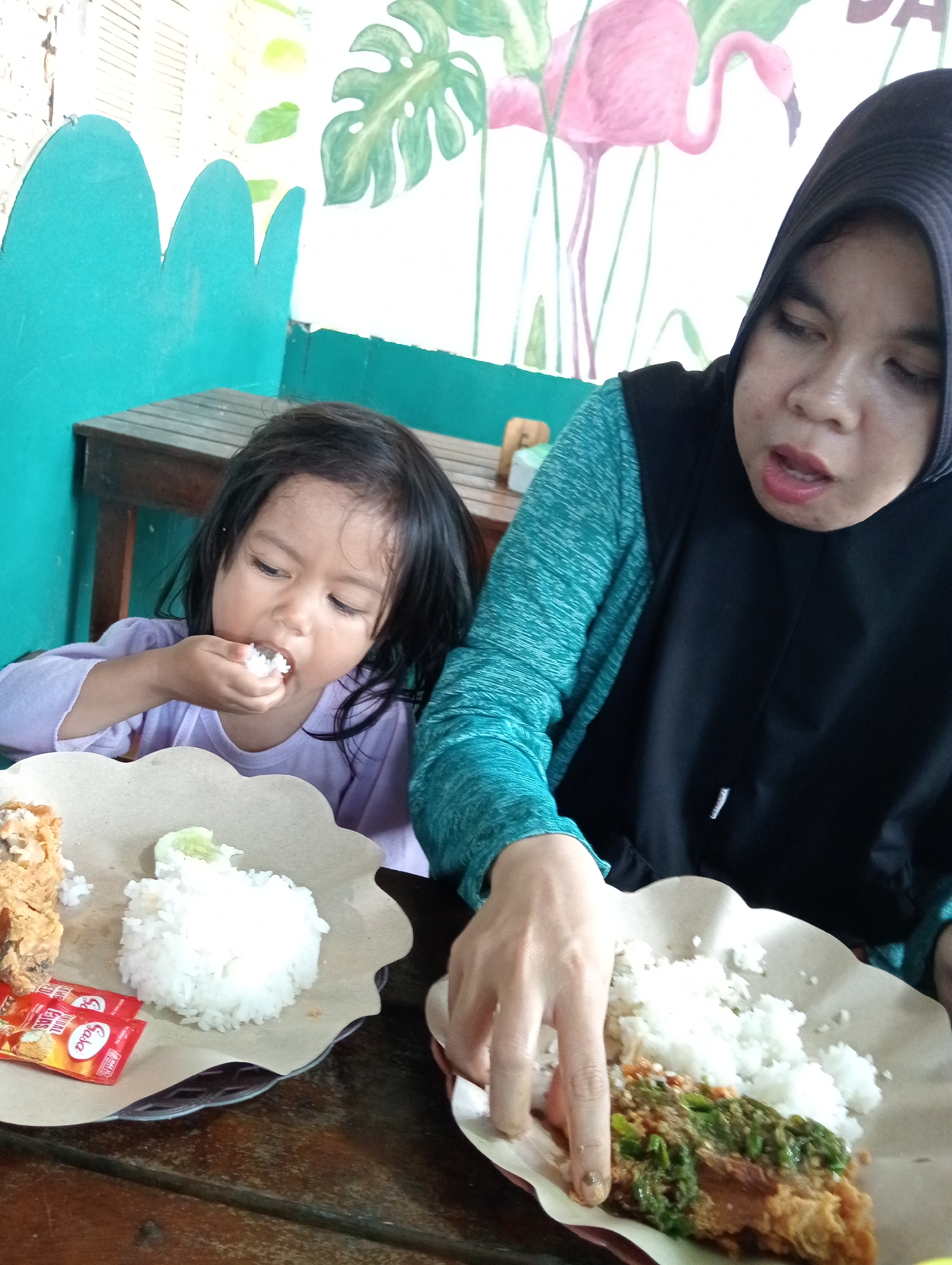 A mother and daughter are eating at a table in what appears to be an outdoor dining area. The daughter, on the left, is a young girl with dark hair wearing a light purple long-sleeved shirt. She is holding a spoonful of rice to her mouth. In front of her is a plate with white rice, a piece of fried chicken, and a slice of cucumber. The mother, on the right, is wearing a black hijab and a teal and black patterned long-sleeved shirt. Her hands are visible as she reaches towards her plate, which also contains rice and fried chicken with a green chili sauce. The background features a brightly colored mural with stylized green leaves and pink flamingos. The table is made of wood, and the overall setting suggests a casual dining establishment, possibly in the city of Campago, Indonesia, given the context.