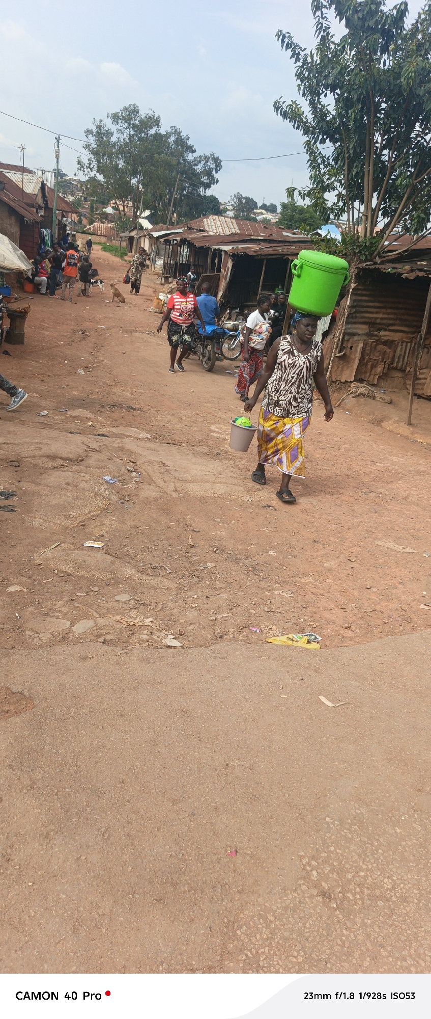 In Barikin Gidar Kanat, Nigeria, a woman walks down a dirt road carrying a large green plastic container on her head and a gray bucket in her right hand. The bucket appears to contain a green object. She is wearing a patterned sleeveless dress and a yellow and purple patterned skirt. Behind her, several other people are visible in the street. To the left of the woman, a motorcycle with two people on it is stopped. The buildings lining the street are simple structures with corrugated metal roofs. The ground is dry and dusty, with scattered debris. The sky is overcast.