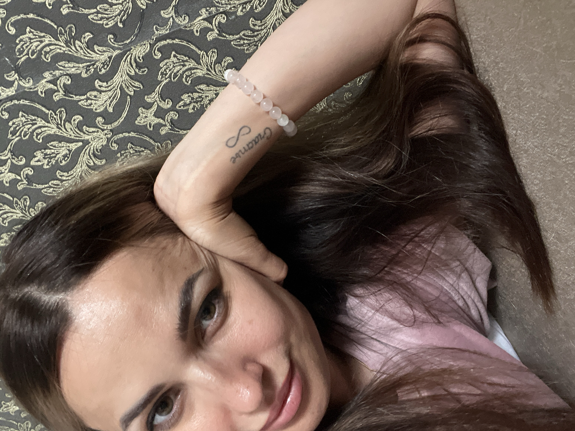 A woman with long, dark brown hair rests her head on her hand, looking at the camera with a slight smile. She is wearing a pink shirt and a bracelet with light pink beads on her left wrist. A tattoo of an infinity symbol and script is visible on her wrist. The background is a dark, textured wallpaper with a gold floral pattern. The scene appears to be indoors.