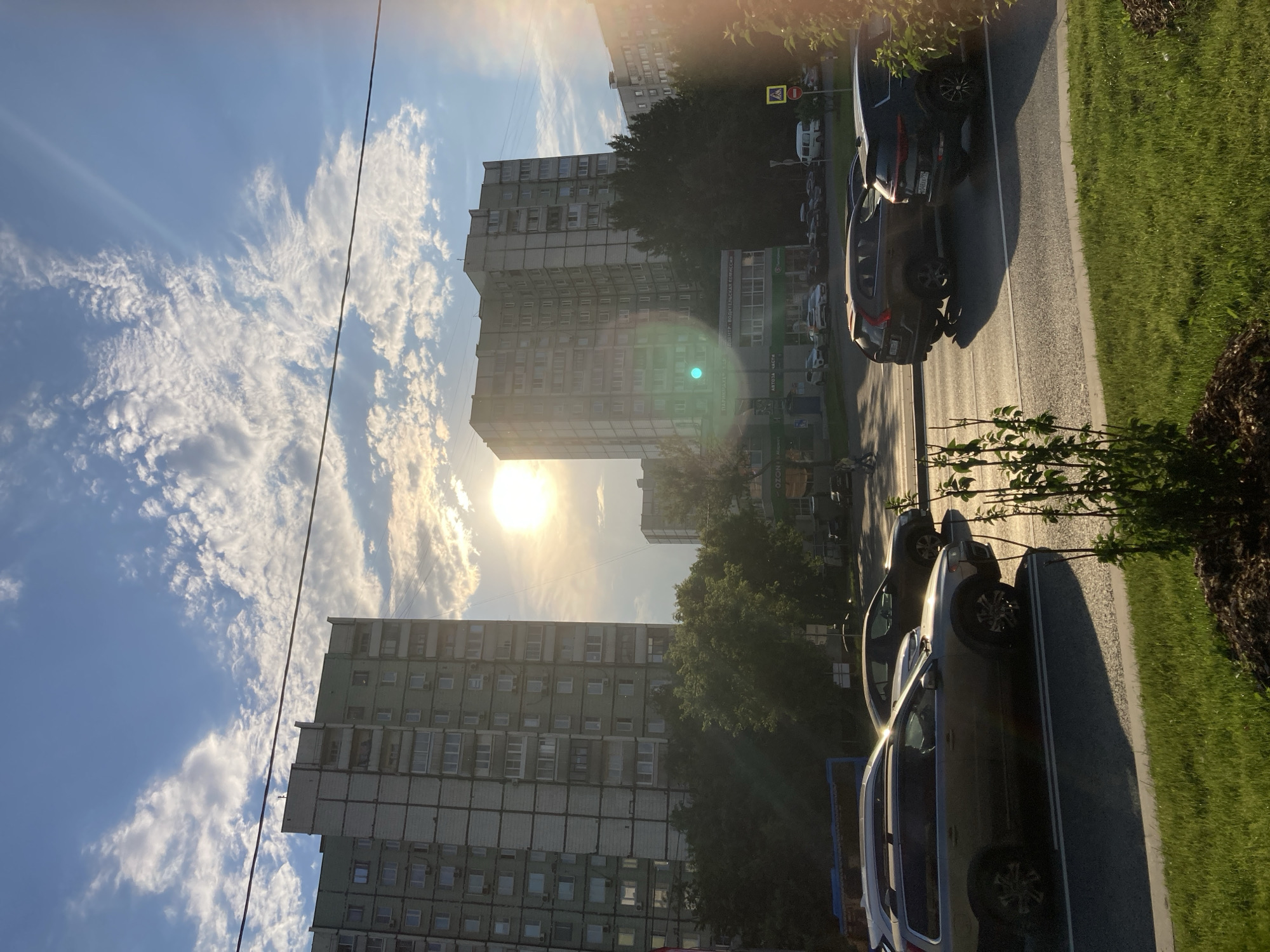 The image captures a view of a street in Moscow, Russia, during what appears to be late afternoon or early evening, given the low angle of the sun and the golden light illuminating the scene. Two large, multi-story residential buildings, typical of Soviet-era architecture, dominate the background. These buildings are light-colored, possibly concrete or stucco, with numerous windows. Trees and greenery are visible between the buildings and in the foreground.
In the middle ground, a busy street is depicted with several cars in motion and parked. The cars are varied, including SUVs and sedans. Some buildings with storefronts and signage are also visible along the street. A pedestrian or two can be seen walking on the sidewalk.
The sky is a vibrant blue, dotted with fluffy white clouds, and the sun is shining brightly, creating lens flare and casting long shadows across the street and grass. The foreground features a well-maintained grassy verge and a small, leafy plant. A power line stretches diagonally across the upper portion of the frame. The overall impression is one of an urban residential area bathed in the warm light of the setting sun.