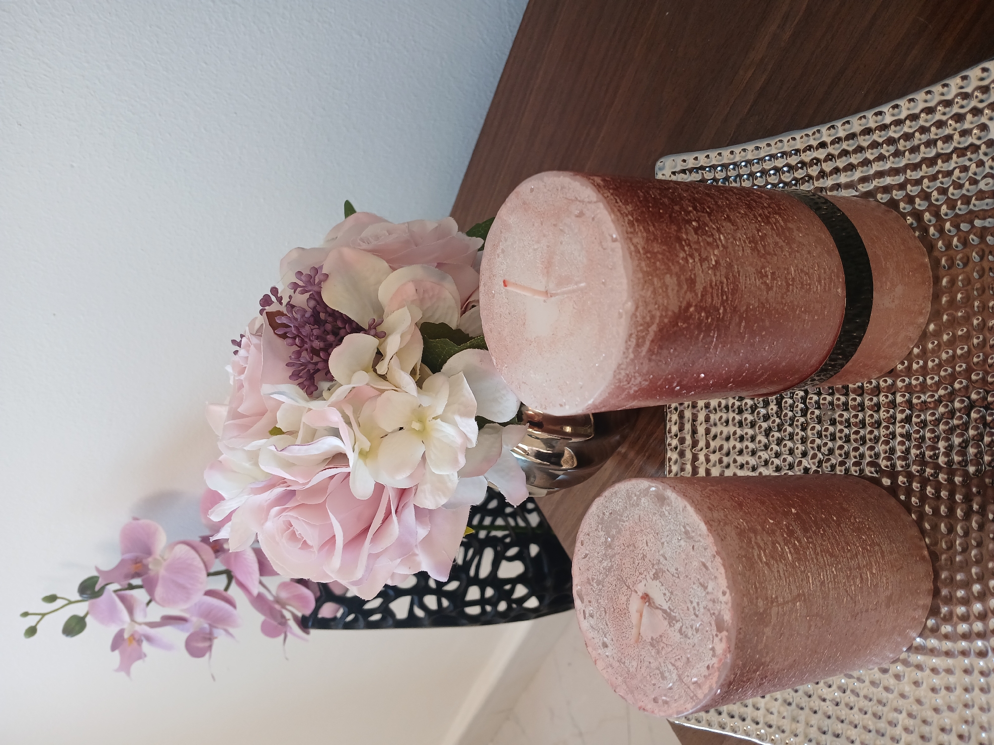 The image shows a still life arrangement, seemingly indoors. On a dark brown wooden surface, there's a bouquet of artificial flowers, predominantly pink and white, with hints of purple. The flowers are arranged in a black geometric vase. Next to the flowers are two cylindrical, light pink candles with wicks, placed on a textured silver tray. The background is a plain white wall. The composition suggests a decorative setting, possibly in a home. There is no information about the city location.