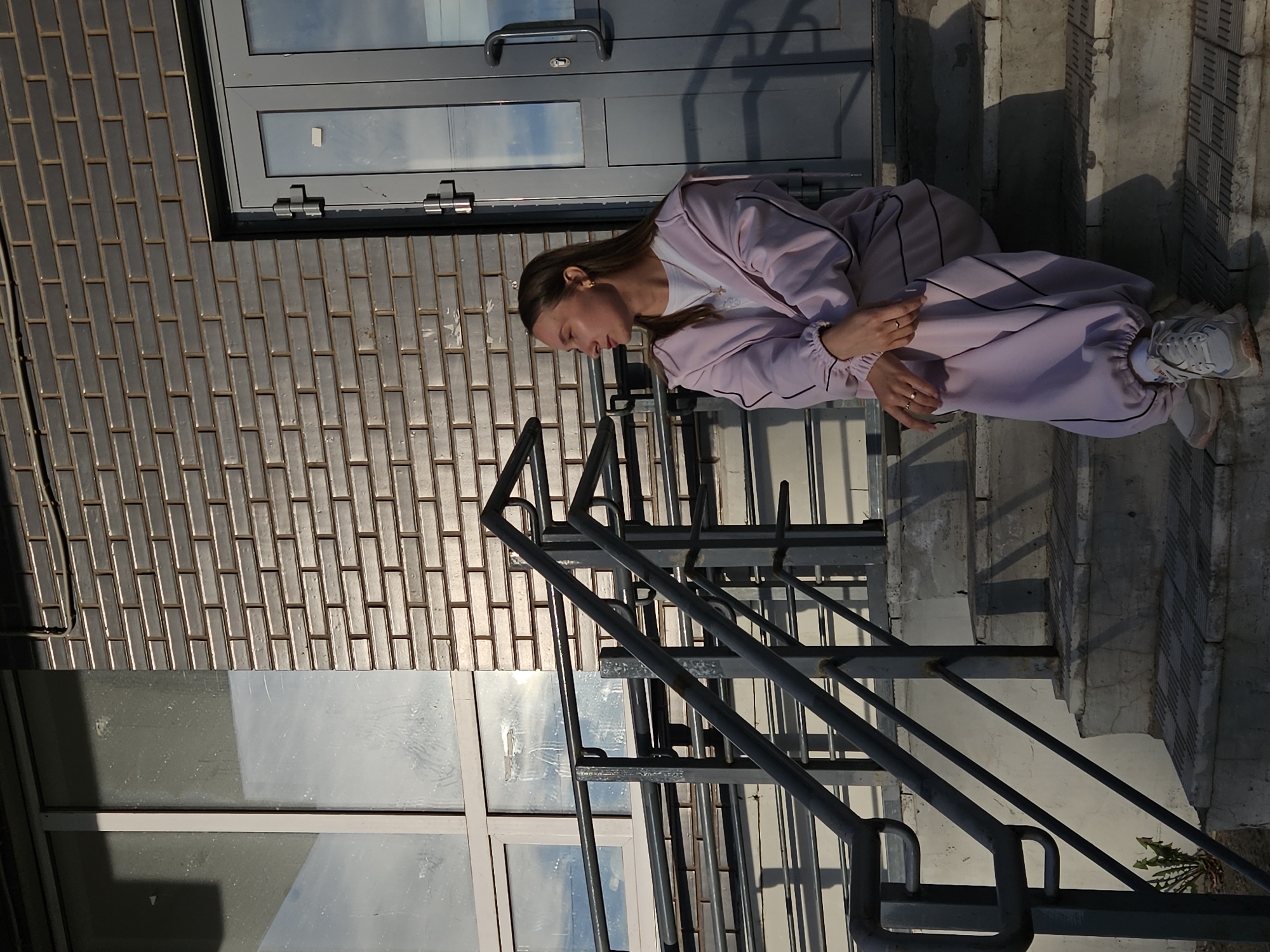A young woman is depicted sitting on concrete steps outside a building in Chaikovsky, Russia. She is positioned upright, facing slightly to her left, with her head bowed and gaze directed downwards, suggesting a contemplative or relaxed state. She has long brown hair and is wearing a light purple tracksuit with dark stripe accents, a white t-shirt, and light-colored sneakers. She has rings on her fingers.
In the foreground, a dark metal railing runs diagonally. Behind the woman, to her left, is a brick wall with horizontal rectangular bricks. To her right, a light-colored door with a window and a handle is visible. Below the brick wall and door, a large window reflects the bright, clear sky.
The scene is brightly lit by natural sunlight, casting sharp shadows from the railing and the woman, indicating a sunny day, likely during the morning or late afternoon. A small green plant grows at the bottom right of the concrete steps. No visible text is present in the image.
