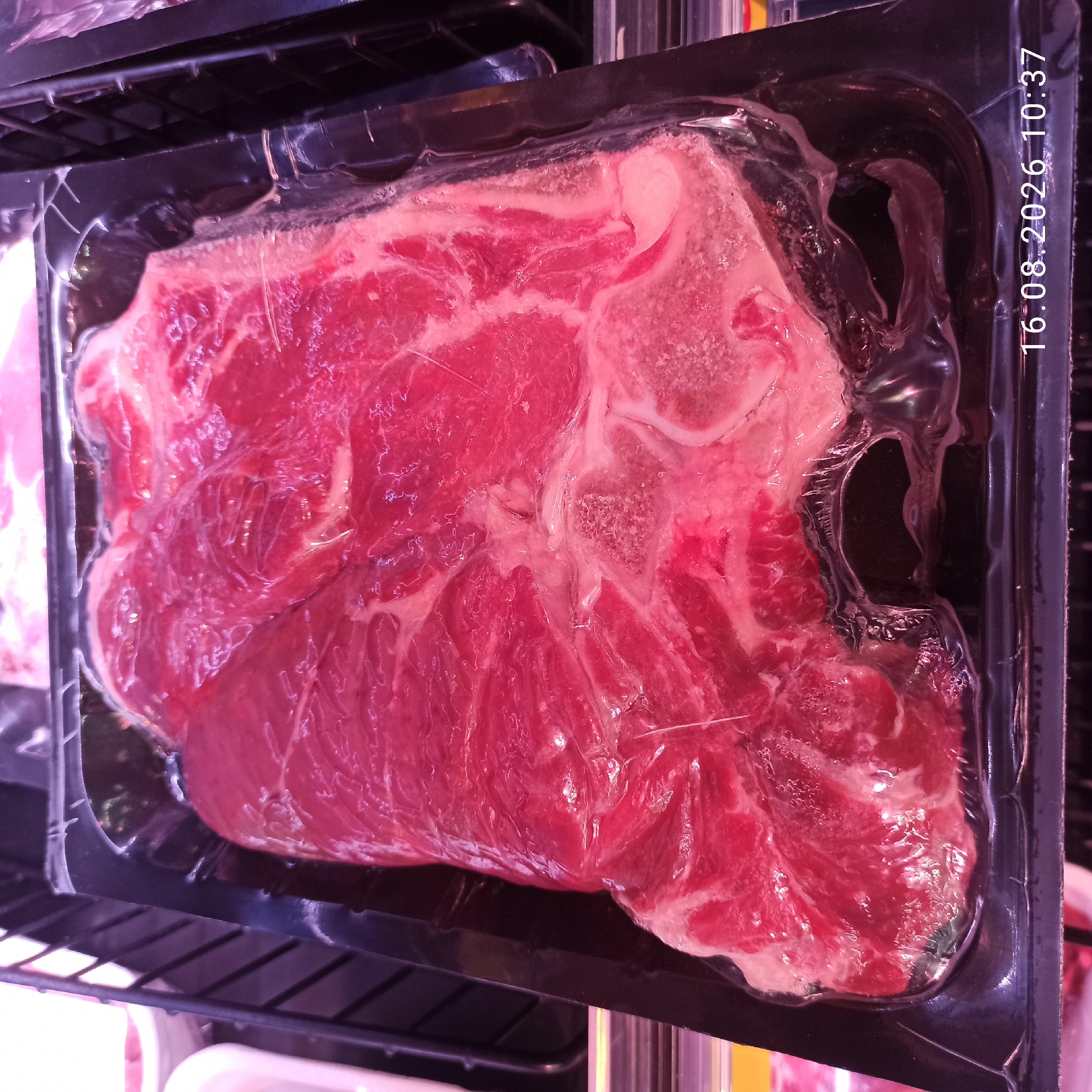 The image shows a vacuum-sealed package of raw meat, likely beef, resting in a black plastic tray. The meat is a deep red color, with visible marbling and some bone fragments. The plastic packaging is transparent, allowing a clear view of the meat's texture and marbling. The tray appears to be part of a larger display, possibly in a supermarket or butcher shop, suggested by the blurred background which hints at shelves and other products. The lighting in the scene is somewhat dim and artificial, casting a slight pinkish hue over the meat. A timestamp, "16.08.2026 10:37", is visible in the lower right corner of the image, indicating a specific date and time. The image does not depict any people or any specific location in the city of Terlizzi, Italy; it is a close-up of a packaged food item.