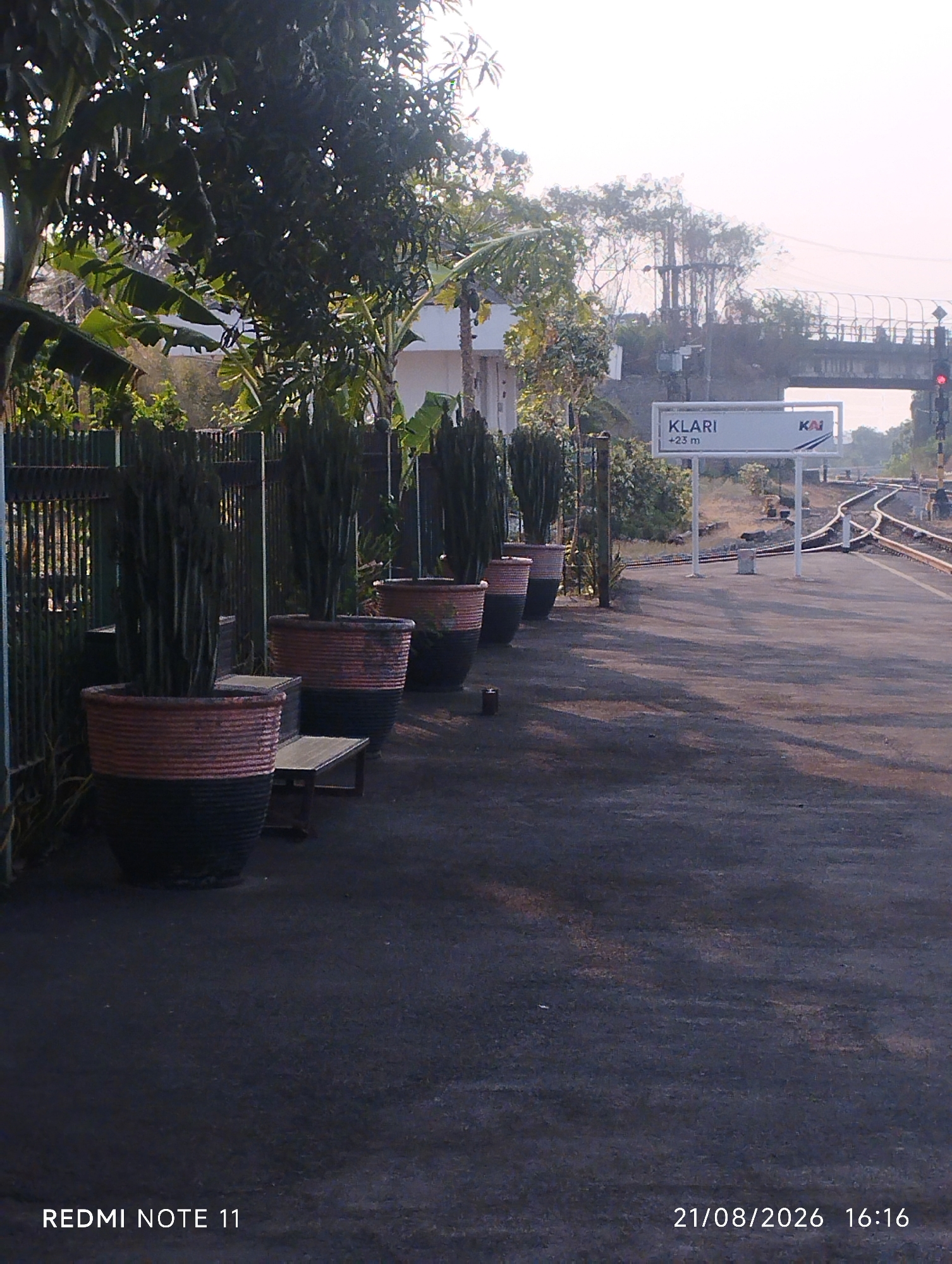 The image shows a train station platform at Klari, Karawang, Indonesia, with a sign that reads "KLARI +23 m KAI". The platform is paved with dark asphalt and has train tracks running along the right side of the image. In the foreground, there are several large potted plants with tall, dark green succulents. A metal fence lines the left side of the platform, behind which are trees and some buildings. In the background, an overpass and traffic signals are visible, along with the railway tracks continuing into the distance. The lighting suggests it is late afternoon, with long shadows cast across the platform. The bottom left corner displays "REDMI NOTE 11" and the bottom right corner shows "21/08/2026 16:16", indicating the device and date/time the photo was taken. There are no people or specific actions depicted in the image.