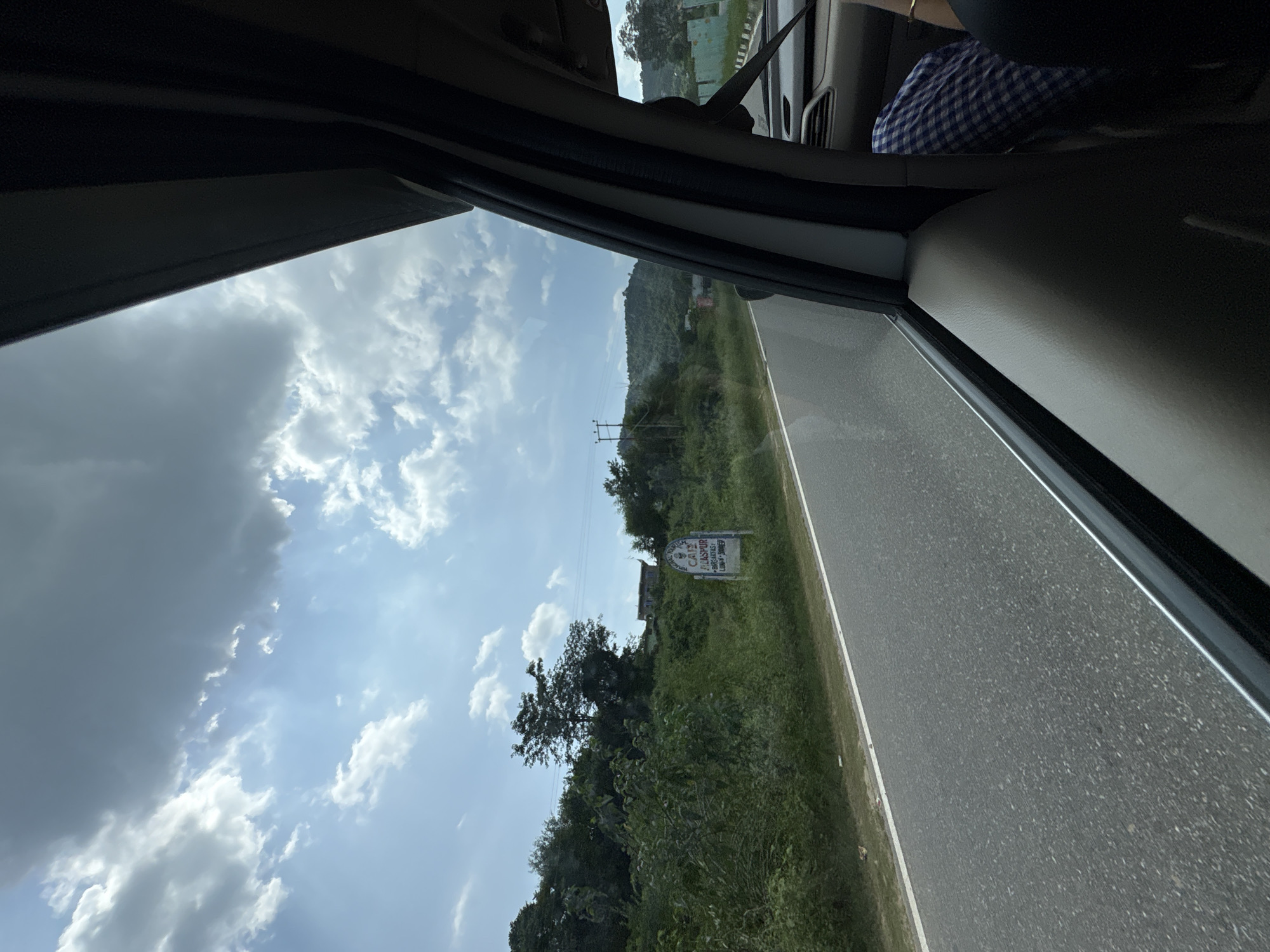 The image depicts a roadside view from inside a vehicle, likely in or around Bilaspur, India, judging by the sign visible on the side of the road. The sky is partly cloudy, with a mix of bright sunlight and dark, heavy clouds. A paved road stretches into the distance, bordered by lush green vegetation and trees. In the middle ground, a sign with text is visible, appearing to advertise a "Cafe" and "Bilaspur." Further down the road, electrical poles and distant hills are discernible. The interior of the vehicle is partially visible, showing a portion of a seat and a person's lap with checkered fabric. The perspective suggests the photo was taken while the vehicle was in motion.