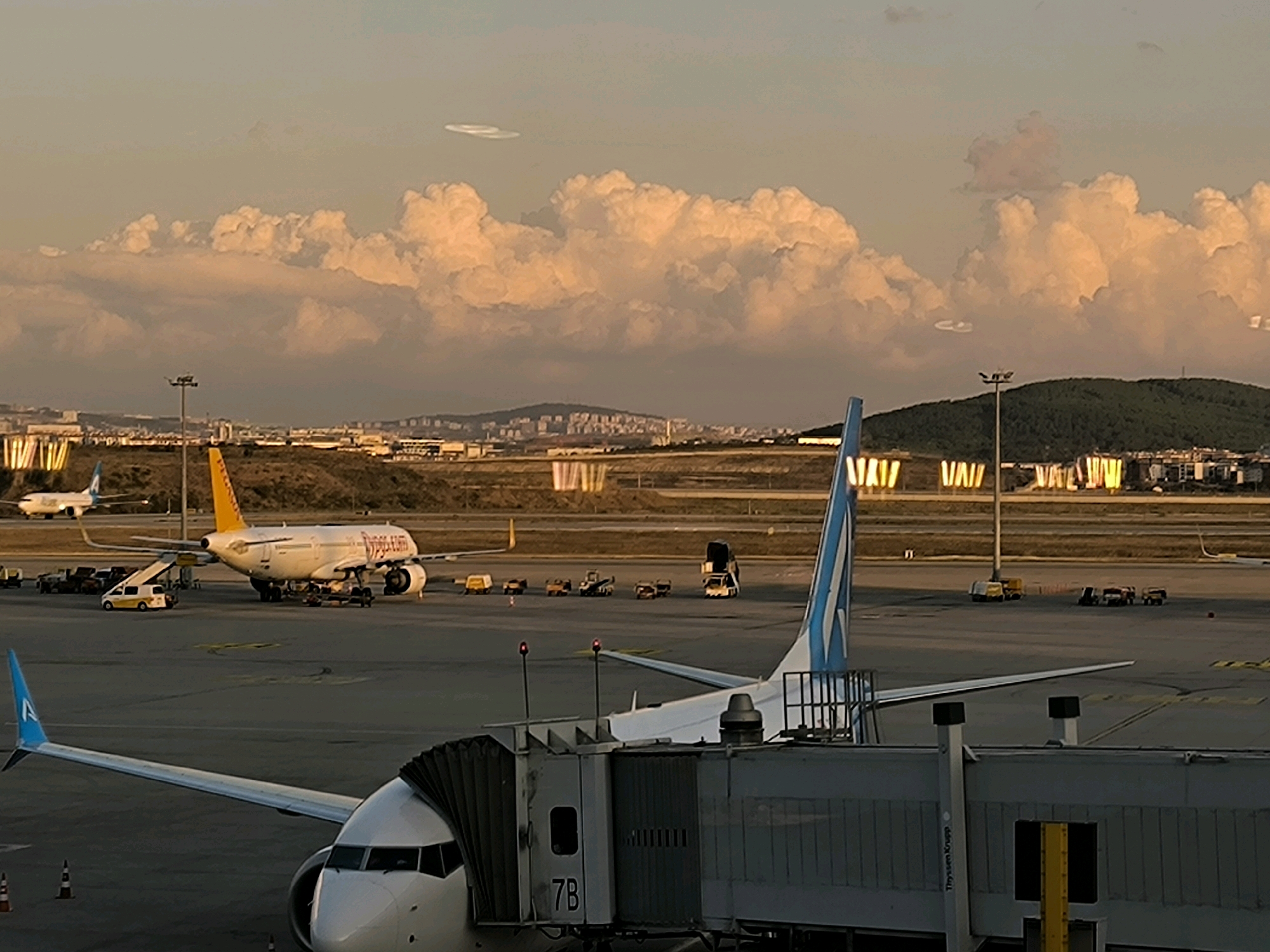 The image depicts an airport tarmac scene at dusk, with a focus on two airplanes. In the foreground, the nose and wing of a white aircraft are visible, connected to a jet bridge labeled "7B". Beyond this, a yellow and white Pegasus Airlines plane is parked, with its engines and tail visible. Several airport service vehicles, including a yellow van and baggage carts, are scattered around the tarmac. In the background, a cityscape with buildings and a green, hilly landscape are visible under a sky filled with dramatic, orange-tinged clouds. The lighting suggests the sun is low on the horizon, casting long shadows. The location is identified as Pendik, Turkey.