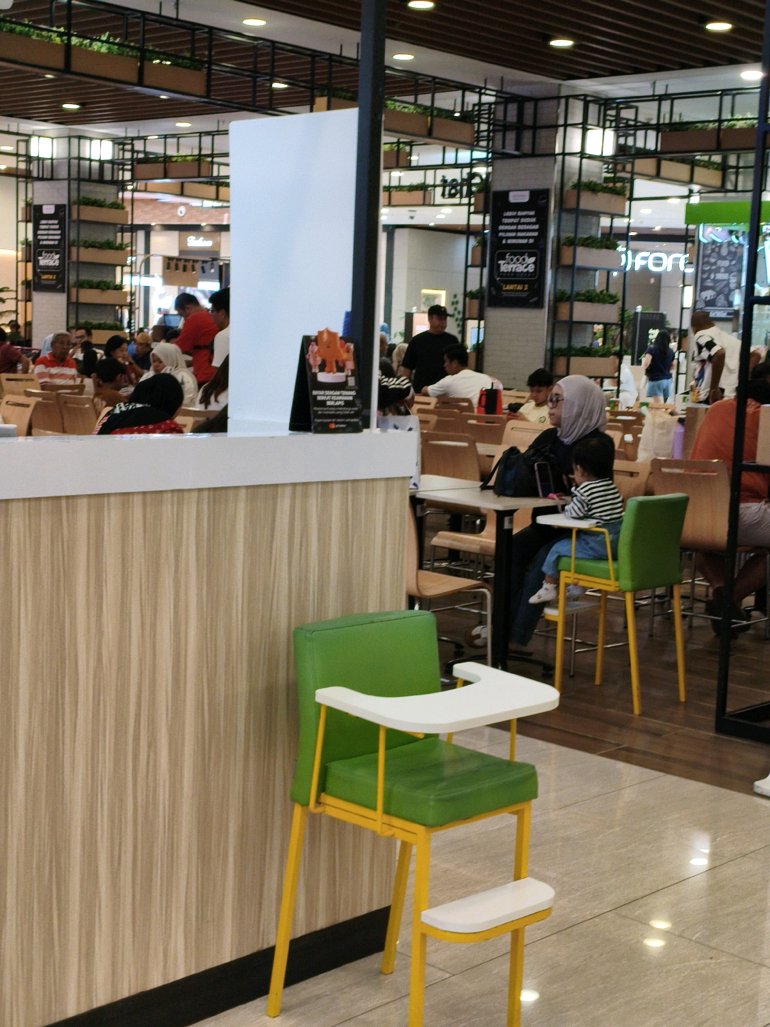 The image depicts a bustling indoor food court, likely located in the city of Babakan Madang, Indonesia. The scene is filled with diners seated at tables and waiting in line. A prominent feature in the foreground is a bright green high chair with yellow legs, positioned near a wooden paneled wall. In the mid-ground, several people are seated at tables, including a woman in a light grey hijab wearing glasses, who is accompanied by a young child in a striped shirt. Behind them, a variety of food stalls and seating arrangements are visible, with people queuing and interacting. The ceiling features a modern design with integrated lighting and decorative shelving containing plants. The overall ambiance suggests a lively and communal dining environment.