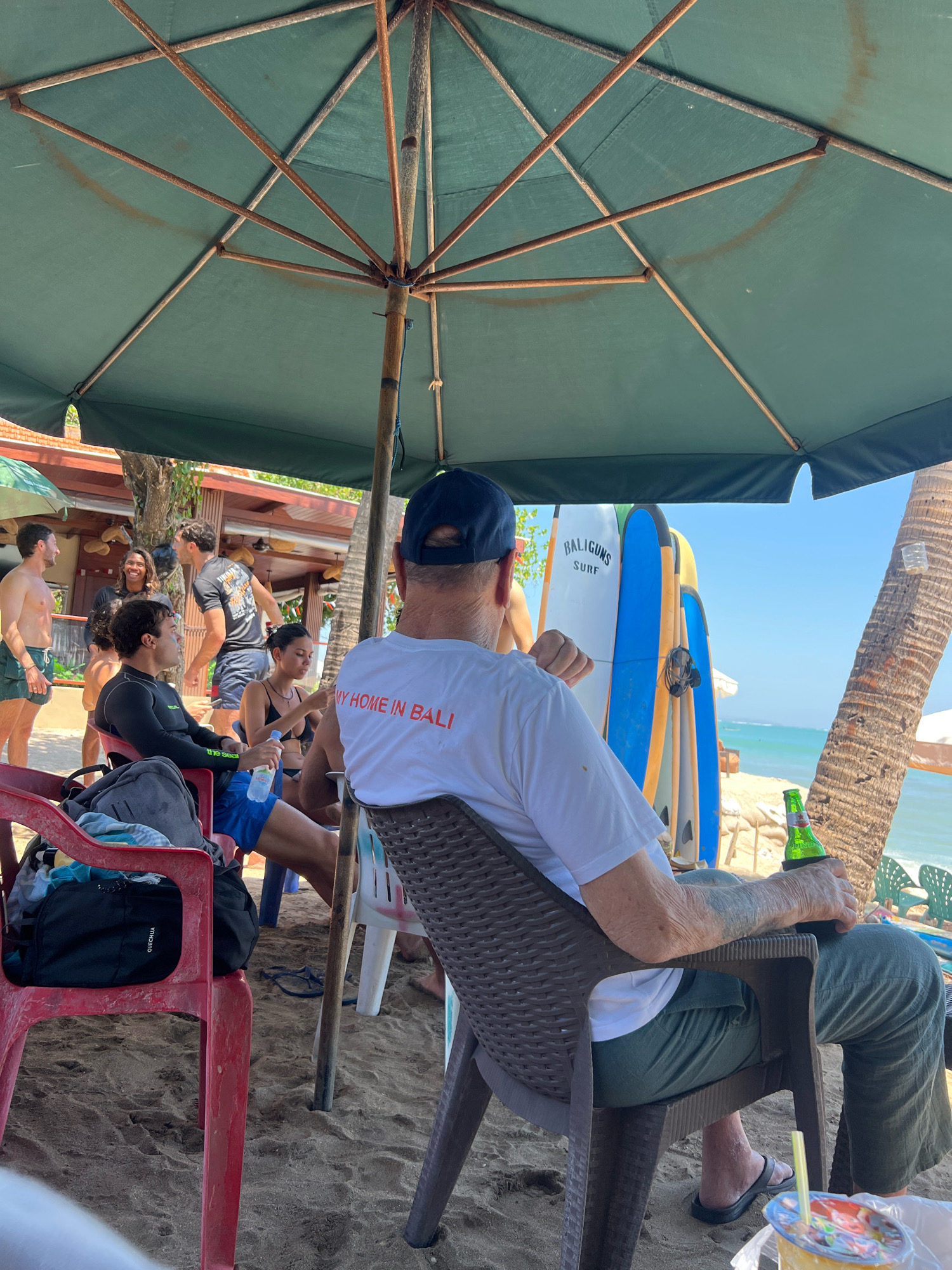 In Kuta, Indonesia, a group of people are gathered on a sandy beach under a large green umbrella. The scene is set near the ocean, with palm trees and a beachside building visible in the background.
A man in the foreground, viewed from behind, wears a white t-shirt with orange text that reads "MY HOME IN BALI" and dark green pants. He is seated in a brown plastic chair, holding a green bottle with a can holder in his right hand. Next to him, to his left, are several surfboards leaning against a support structure. The surfboards have colorful designs, with one white board featuring the text "BALIGUNS SURF."
To the left of the man in the foreground, other individuals are seated on red and white plastic chairs, also on the sand. One person wears a black wetsuit top and blue shorts, while another wears a black bikini. Several other people stand and converse in the mid-ground, some appearing to be tourists.
The environment is characteristic of a beachside establishment in Kuta, with beach chairs, surfboards, and an umbrella providing shade. The ocean is visible in the distance, and the overall atmosphere suggests a relaxed, tropical setting.