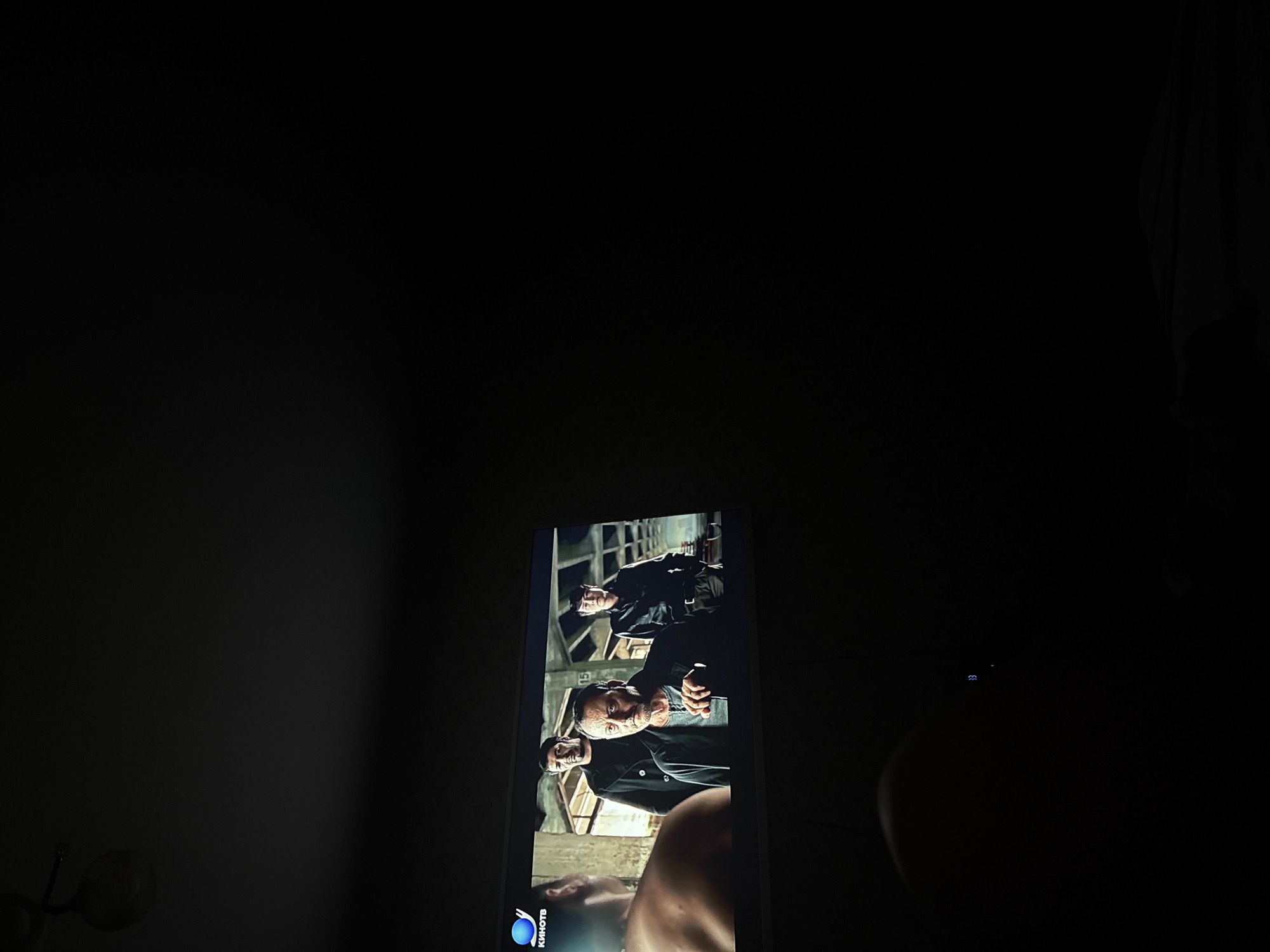 A cell phone displays a scene with three men in dark clothing standing under what appears to be an open-air structure with metal beams. A portion of a bare-skinned person is visible at the bottom of the screen. The text "КИНОТВ" is displayed in the lower-left corner of the screen. The phone sits in a dark room with a wall outlet visible on the left. A curtain or fabric is visible on the right side of the frame. There is nothing to identify the location in Mytishchi, Russia.