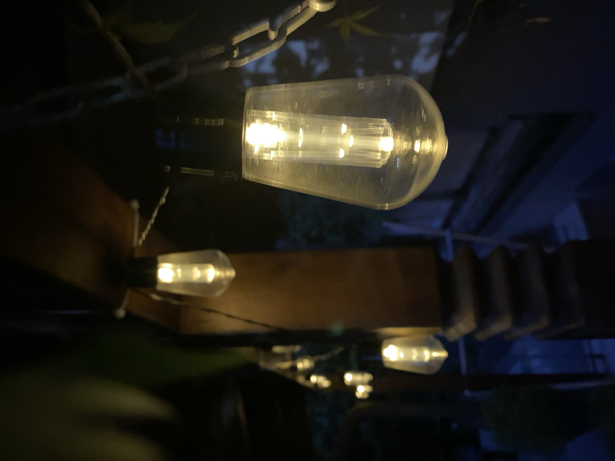 The image captures a string of outdoor decorative lights, likely LED Edison bulbs, illuminating a dark, possibly evening or nighttime, environment. The lights are suspended from a wooden structure and a metal chain, casting a warm, yellow glow. The bulbs themselves are transparent with visible internal elements that emit the light. In the foreground, one bulb is in sharp focus, showing its textured glass and the detailed internal light source. Behind it, a series of other bulbs are visible, receding into a soft blur, with some appearing to be attached to a dark pillar or column. Green foliage is present in the background, suggesting an outdoor setting like a garden, patio, or balcony. The overall impression is one of ambiance and gentle illumination. The image was taken in Purkersdorf, Austria.