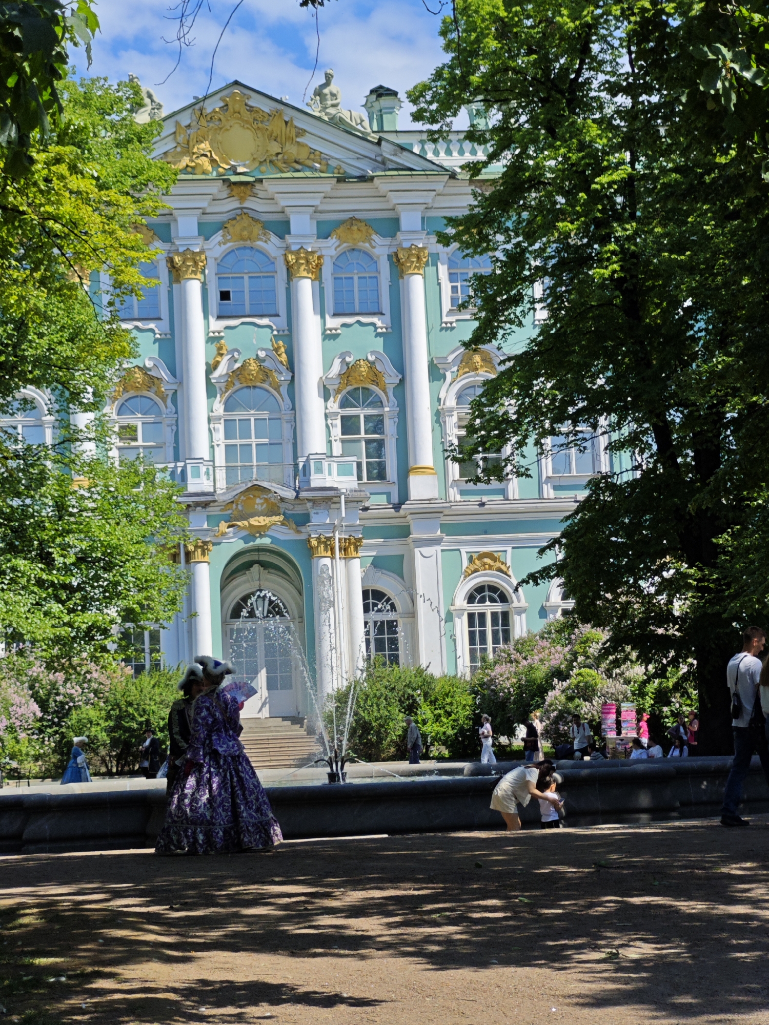 The image depicts the exterior of the Catherine Palace in Pushkin, Saint Petersburg, Russia. The palace is characterized by its striking turquoise facade, adorned with white columns and ornate gold detailing on the pediments and window frames. Lush green trees frame the scene, with their branches casting dappled shadows on the sunlit ground.
In the foreground, a stone fountain serves as a central element. Several people are visible interacting with the environment. Two individuals in historical costumes, one wearing a voluminous purple gown and the other a darker outfit, stand near the fountain, their backs to the viewer. Another person in a light-colored outfit is bending down to interact with a child near the fountain's edge. Further in the background, other individuals are seen walking and observing the surroundings. The sky above is bright blue with scattered white clouds, indicating a clear day. The overall scene suggests a tourist attraction in a park setting, with visitors enjoying the architectural grandeur and natural beauty.