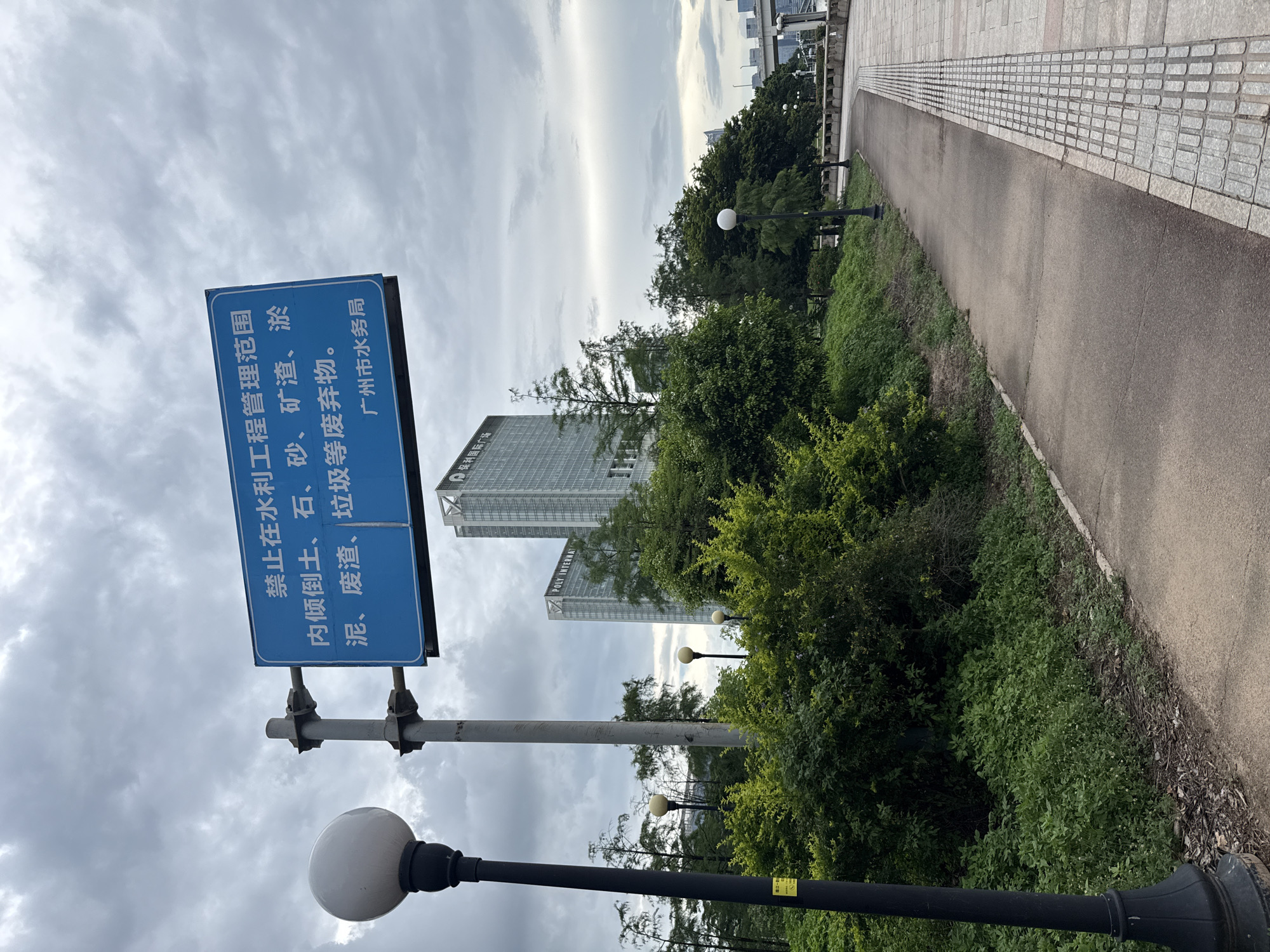 The image shows a paved pathway next to a grassy embankment in Tianhe District, China. A black lamppost with a white spherical light fixture stands on the left side of the pathway. In the background, two modern skyscrapers rise above a lush green canopy of trees and bushes. A blue sign with white Chinese text is affixed to a metal pole in front of the buildings. The sky is overcast with clouds. The pathway is bordered by textured paving stones on the right. The overall scene depicts an urban park or riverside area.