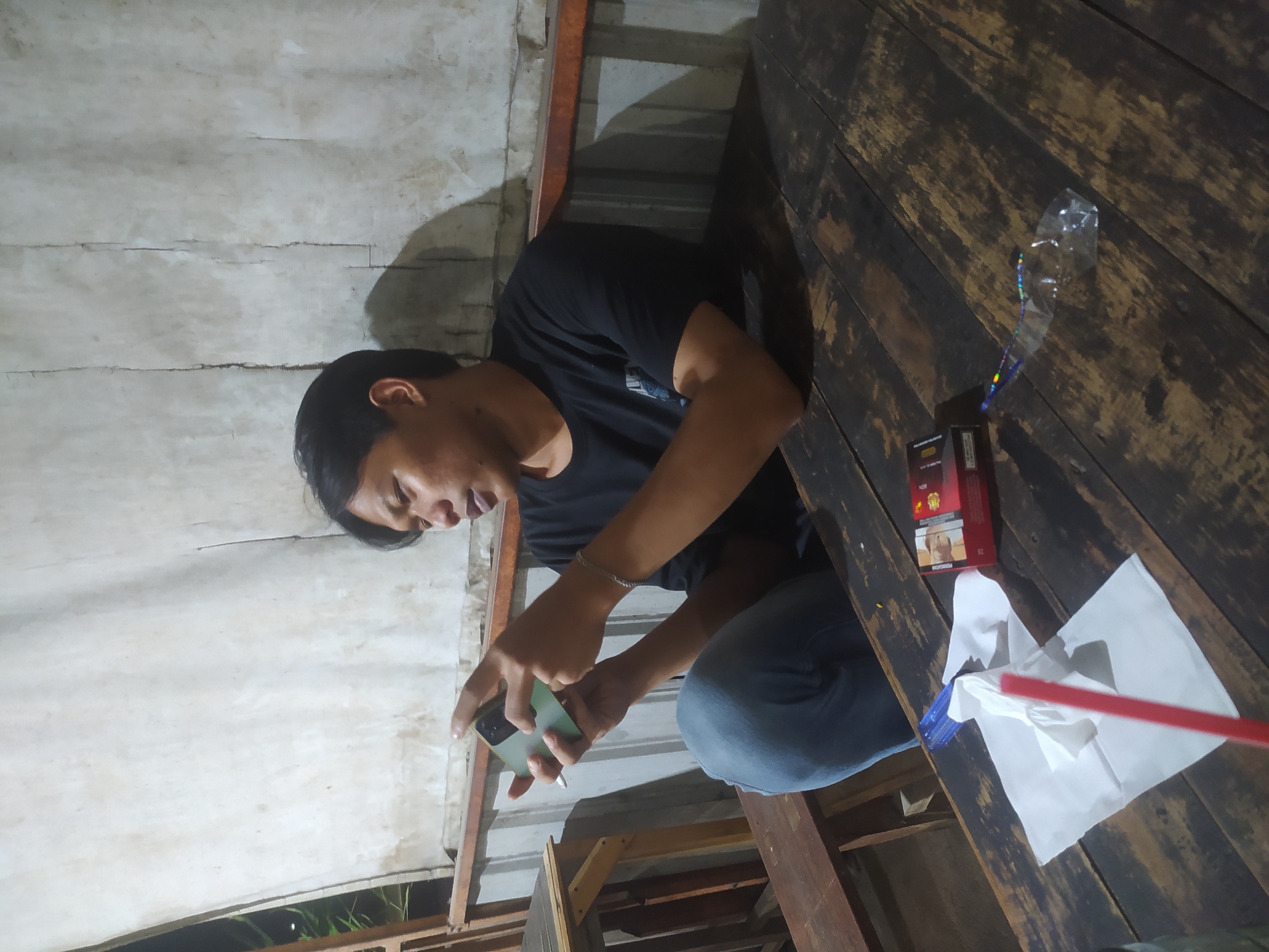 A medium shot captures a young man sitting at a weathered wooden table, engrossed in his smartphone. He wears a black t-shirt and blue jeans. His left hand holds the phone while his right hand rests on the table. To his left, a cigarette pack and a white paper napkin with a red straw are visible on the table. The background shows a textured wall and a railing, suggesting an outdoor or semi-outdoor setting. The scene is illuminated by artificial light, casting shadows on the table and wall. The man appears to be in Tuntang, Indonesia.