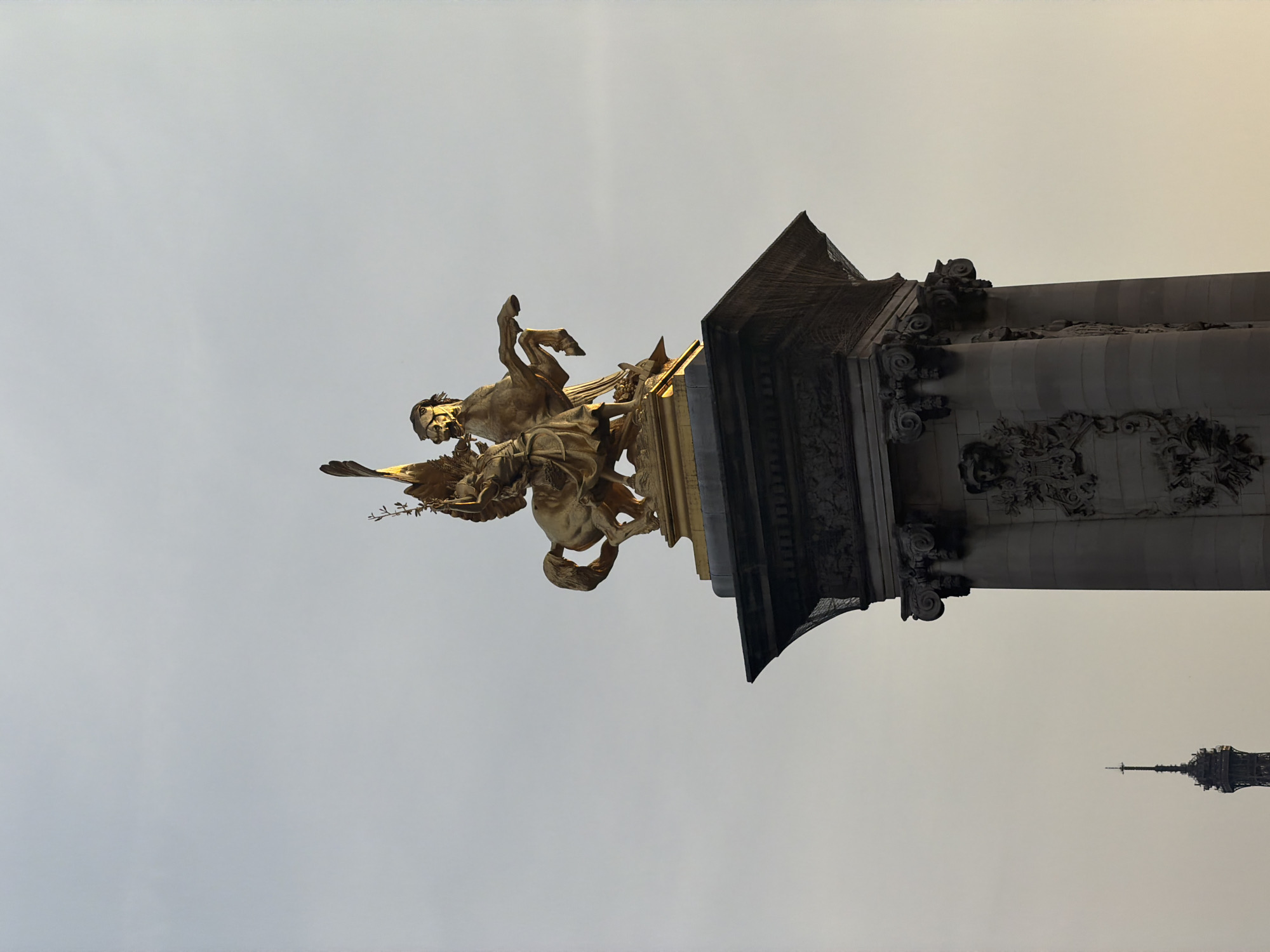 The image depicts a gilded equestrian statue atop a stone pedestal against a pale sky. The statue features a winged figure riding a rearing horse, holding a laurel wreath and a palm branch. The pedestal is ornate, with carved details. In the far distance, the Eiffel Tower is visible, indicating the location is Paris, France, likely near the Pont Alexandre III given the style of the monument. The lighting suggests it is either early morning or late afternoon. The overall scene is a formal, architectural monument.
