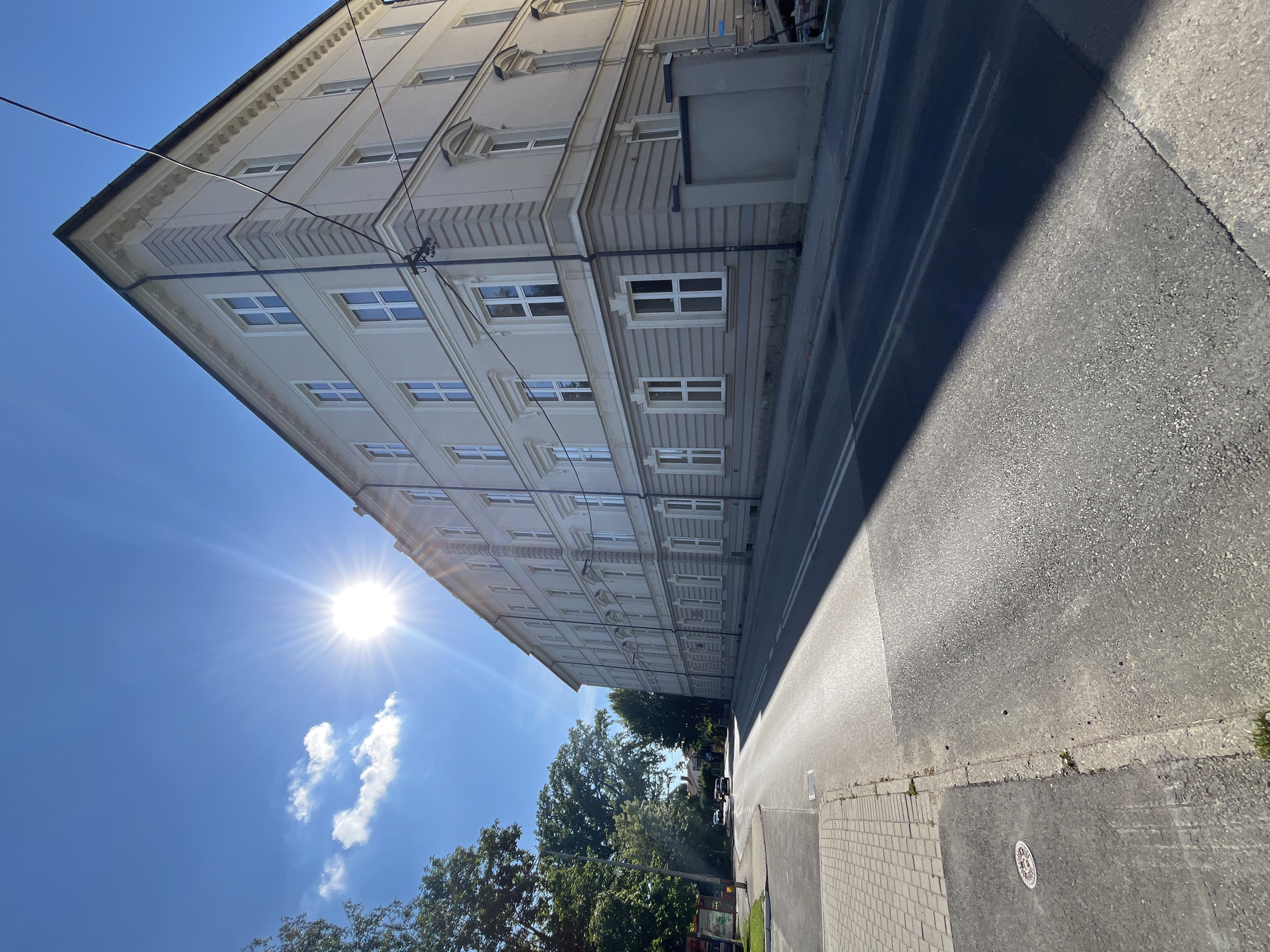 The image shows a street scene in a city on a sunny day. A tall, light-colored building with many windows lines the right side of the street. The building has a classical architectural style with decorative elements around the windows and along the roofline. A bright sun with visible rays shines from the upper left corner of the sky, casting a long shadow across the street. A sidewalk made of pavers runs along the left side of the street. Trees are visible on the left side of the road, and a few cars are driving on the street in the distance. The road itself is paved and appears to have lane markings. The overall impression is a bright, sunny day in an urban environment.