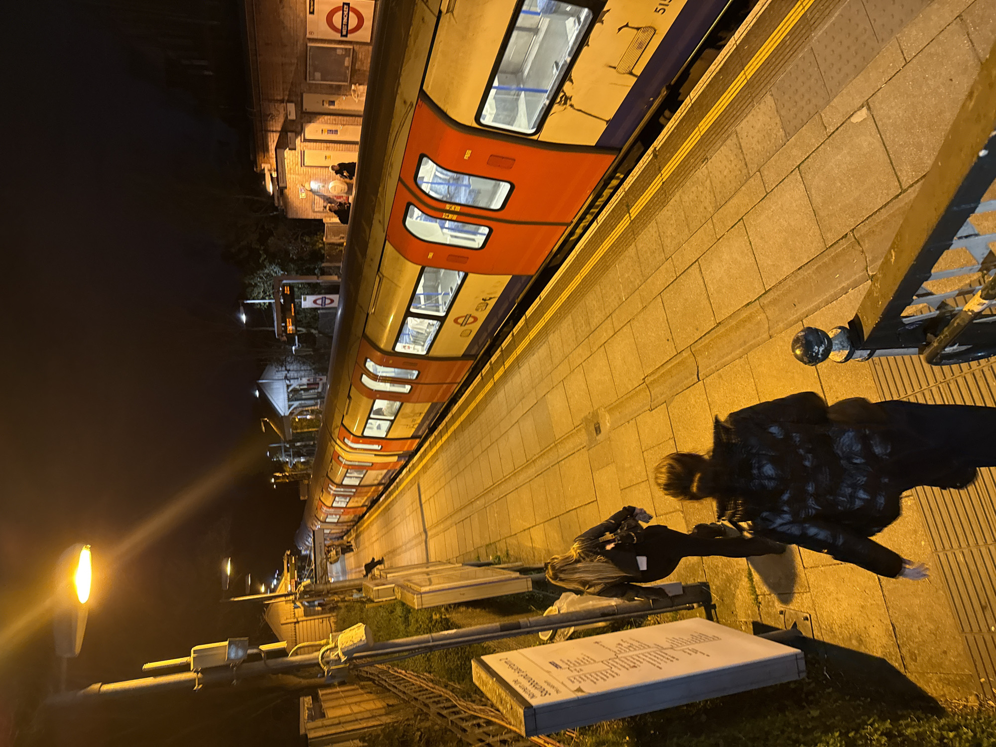 The image shows a platform at the West Finchley Underground station in London, United Kingdom, at night. An orange and white London Underground train is stopped at the platform. Two individuals are walking along the platform, one with a guitar case. A station map is visible, along with platform barriers and lighting. The background shows other station structures and street lighting.