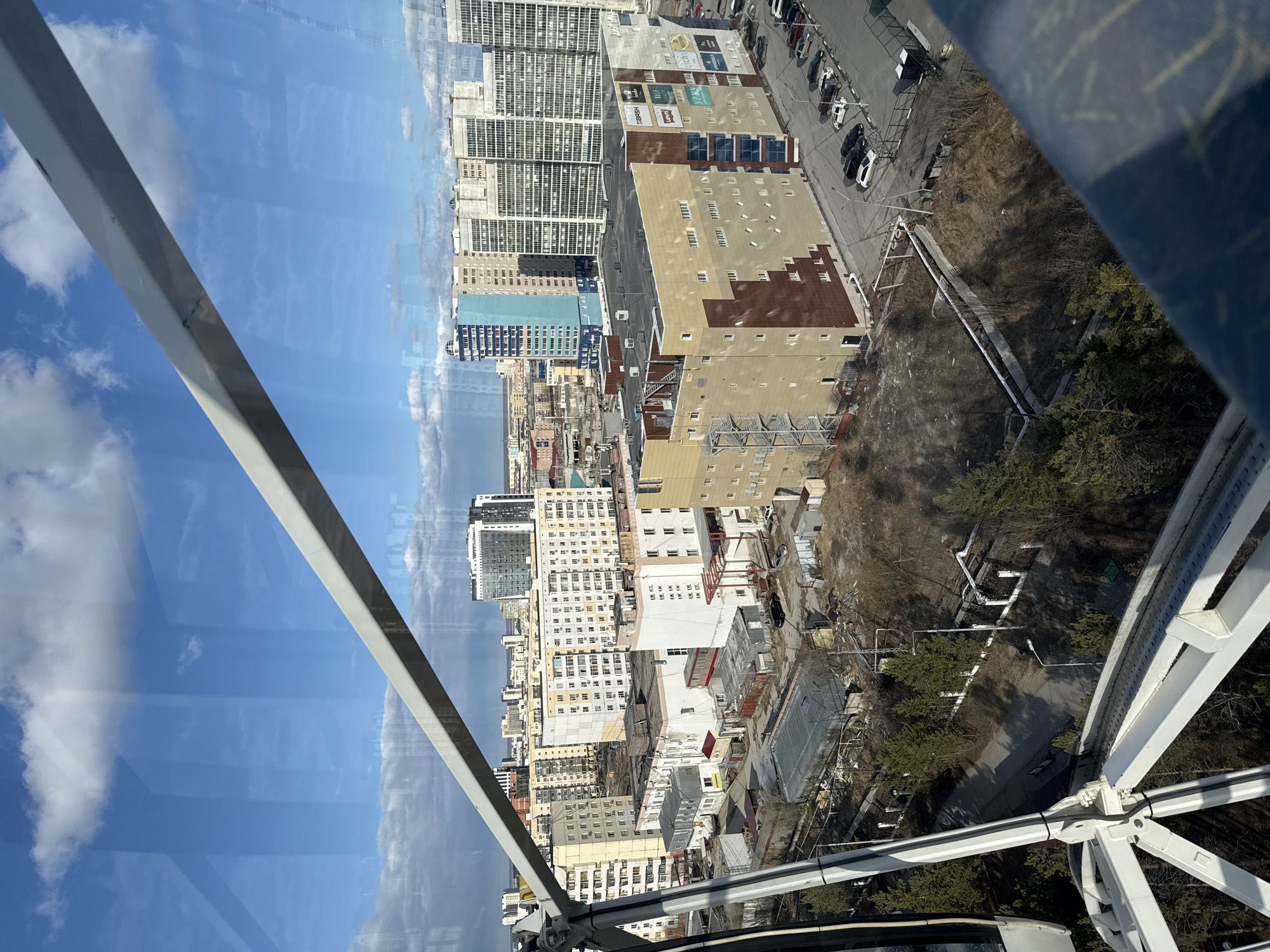 The image depicts an urban scene in Yakutsk, Russia, as viewed from a high vantage point, likely a Ferris wheel. The foreground shows the white framework of the Ferris wheel structure. The midground consists of a dense cityscape featuring buildings of varying heights, styles, and colors, a street with parked cars, and patches of bare ground with scattered trees. The background reveals a bright blue sky with scattered white clouds, partly visible through reflections on glass panels. No people are visible.