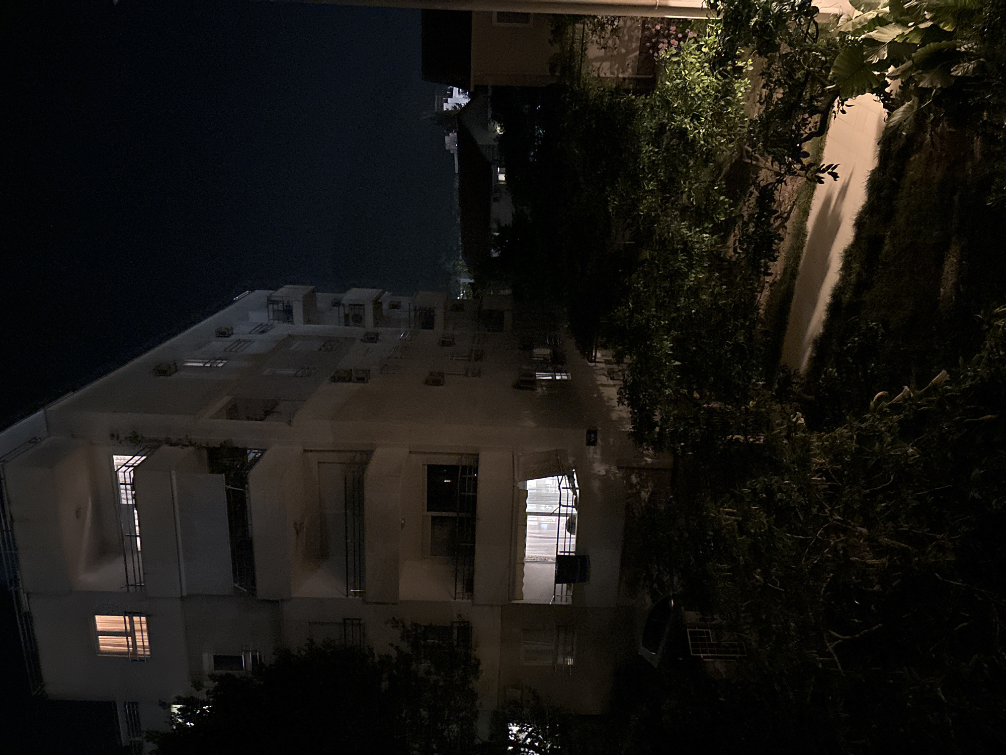 The image is a nighttime photograph of an apartment building in Budva, Montenegro. The building is a light-colored, multi-story structure with several balconies. Lights are visible in some of the windows, indicating occupied apartments. There are trees and foliage in the foreground and around the building, casting shadows in the low light. A pathway is visible through the greenery. In the background, other buildings and a dark sky are visible. The scene is quiet and appears to be residential.