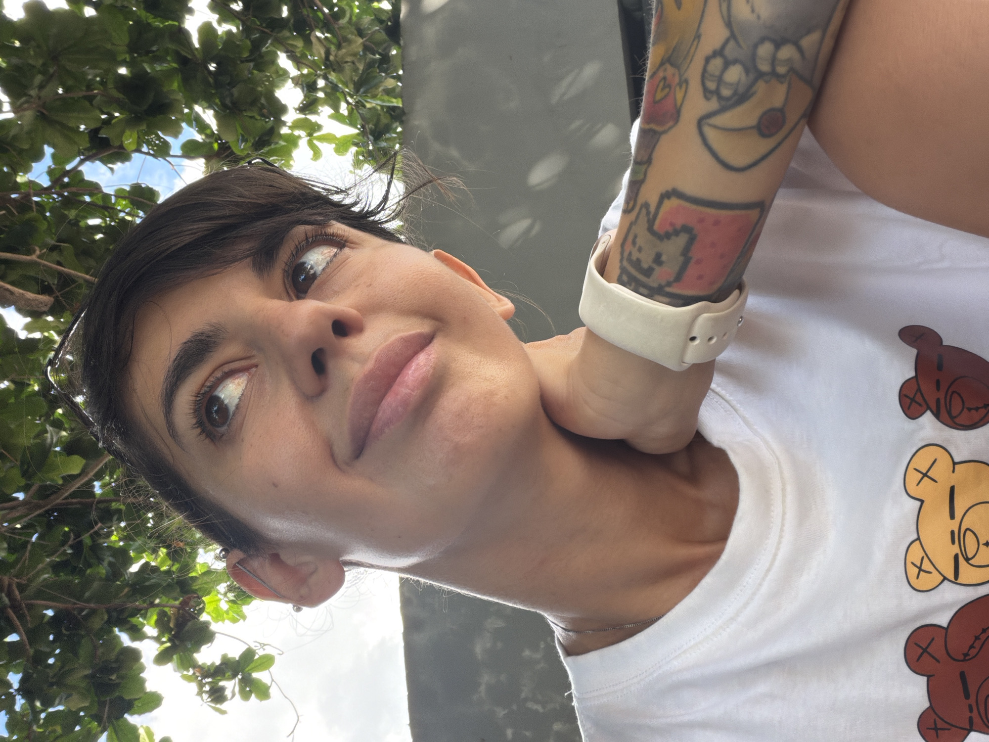 The image shows a close-up of a woman's face and upper body, taken outdoors. She has dark hair, dark eyes, and is wearing a white t-shirt with a design of cartoon teddy bears. Her right arm, which is visible, is covered in tattoos and she is wearing a white smartwatch. She is looking upwards and to her right, with her right hand resting on her neck. Her left arm is not visible. The background consists of green foliage from a tree above and a grey wall behind her, with dappled sunlight creating shadows. The scene suggests the woman is outdoors, possibly in a park or garden, in a warm climate. The presence of a wall, dense foliage, and the style of the building in the background suggest an urban or semi-urban environment. The image was taken at Kuta, Indonesia, suggesting a tropical setting.